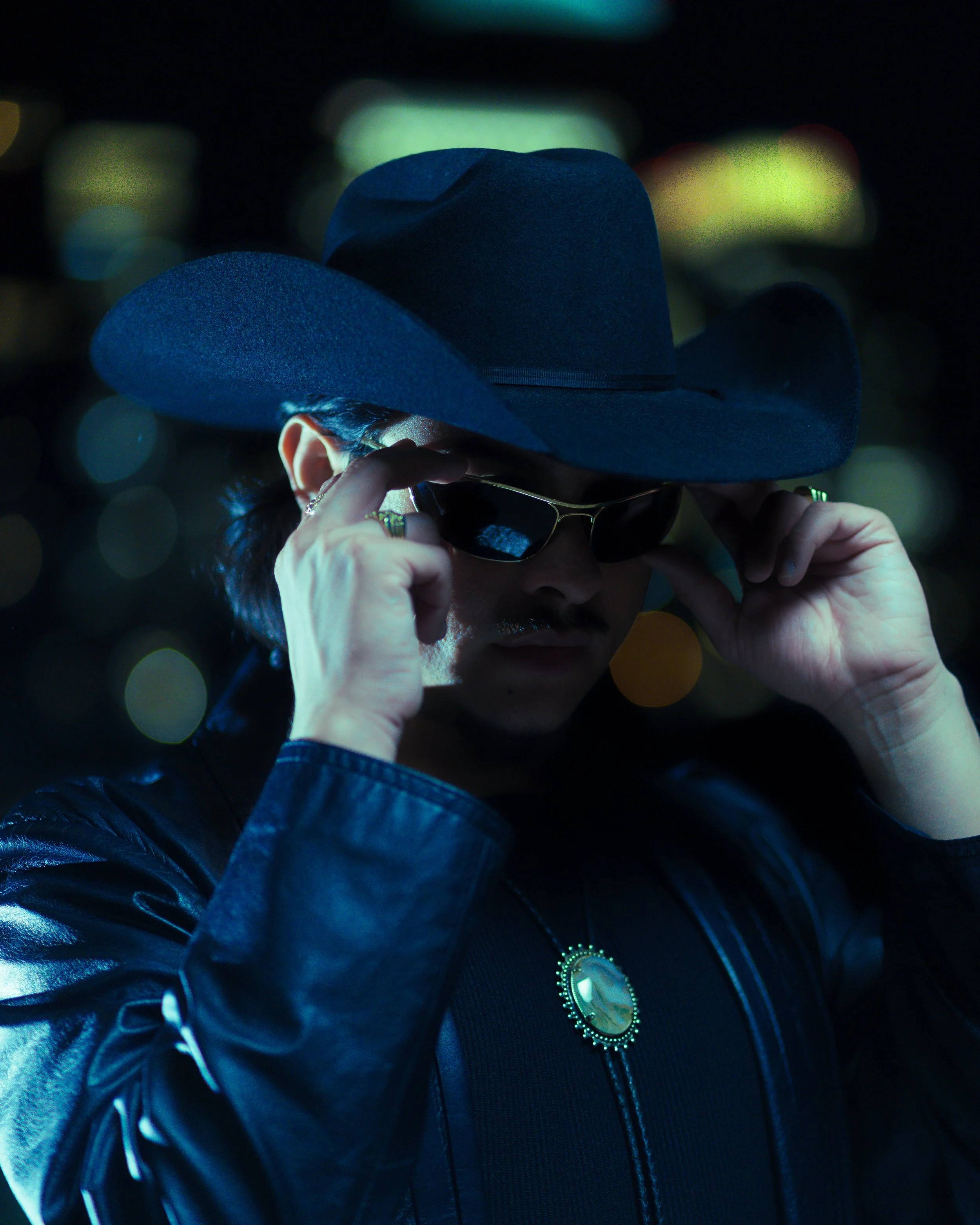 A person wearing a black cowboy hat, dark sunglasses, and a black leather jacket. They are holding their sunglasses with both hands and have a large ornate necklace with a gemstone.