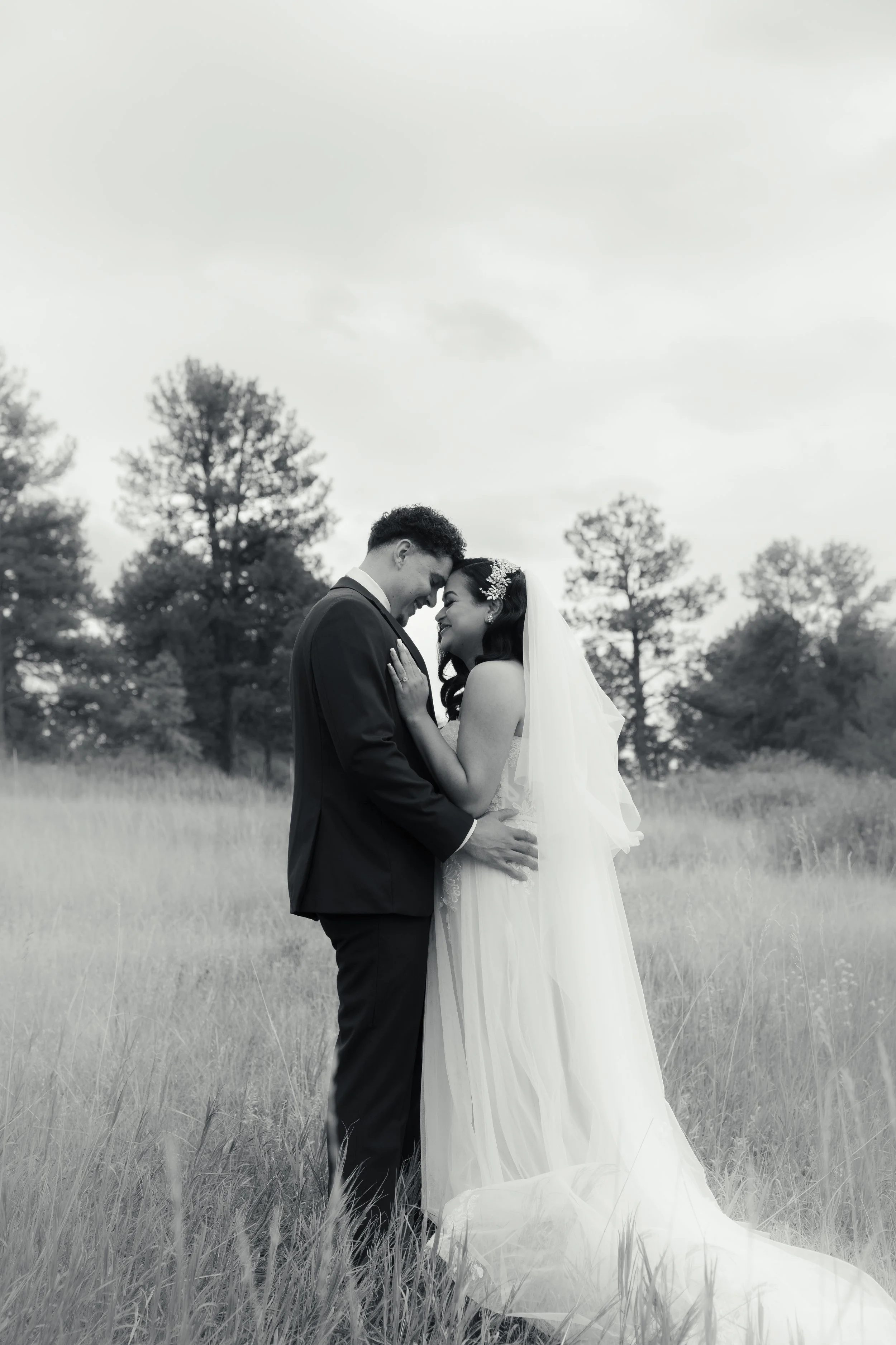Black and white photo of a bride and groom standing close together in a field with trees in the background, embracing and smiling with foreheads touching.