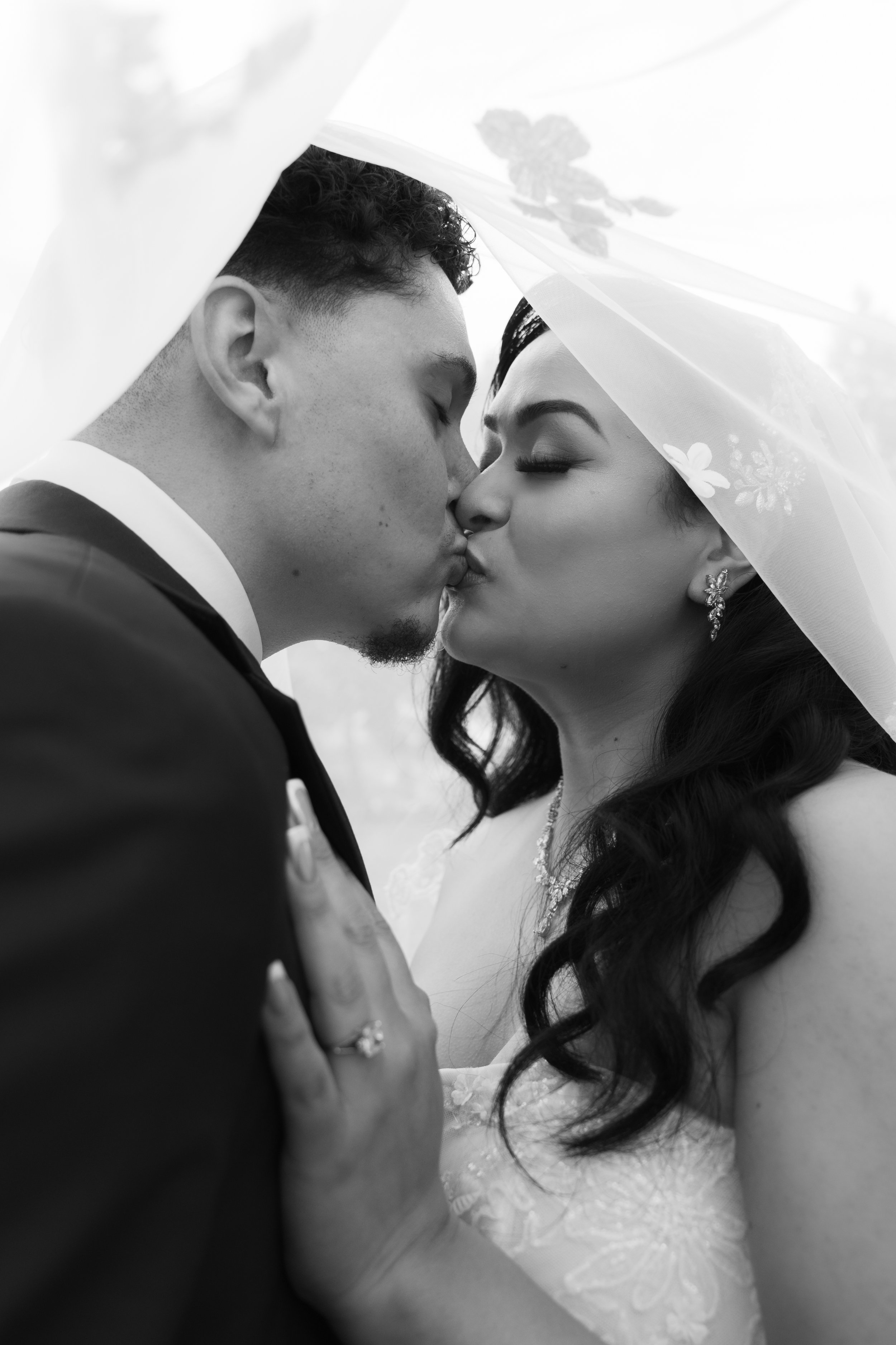 A black and white photograph of a bride and groom sharing a kiss under a veil. The bride has dark hair, earrings, and a necklace. The groom has short curly hair and is dressed in a tuxedo. The bride's hand, showing an engagement ring, is placed on th