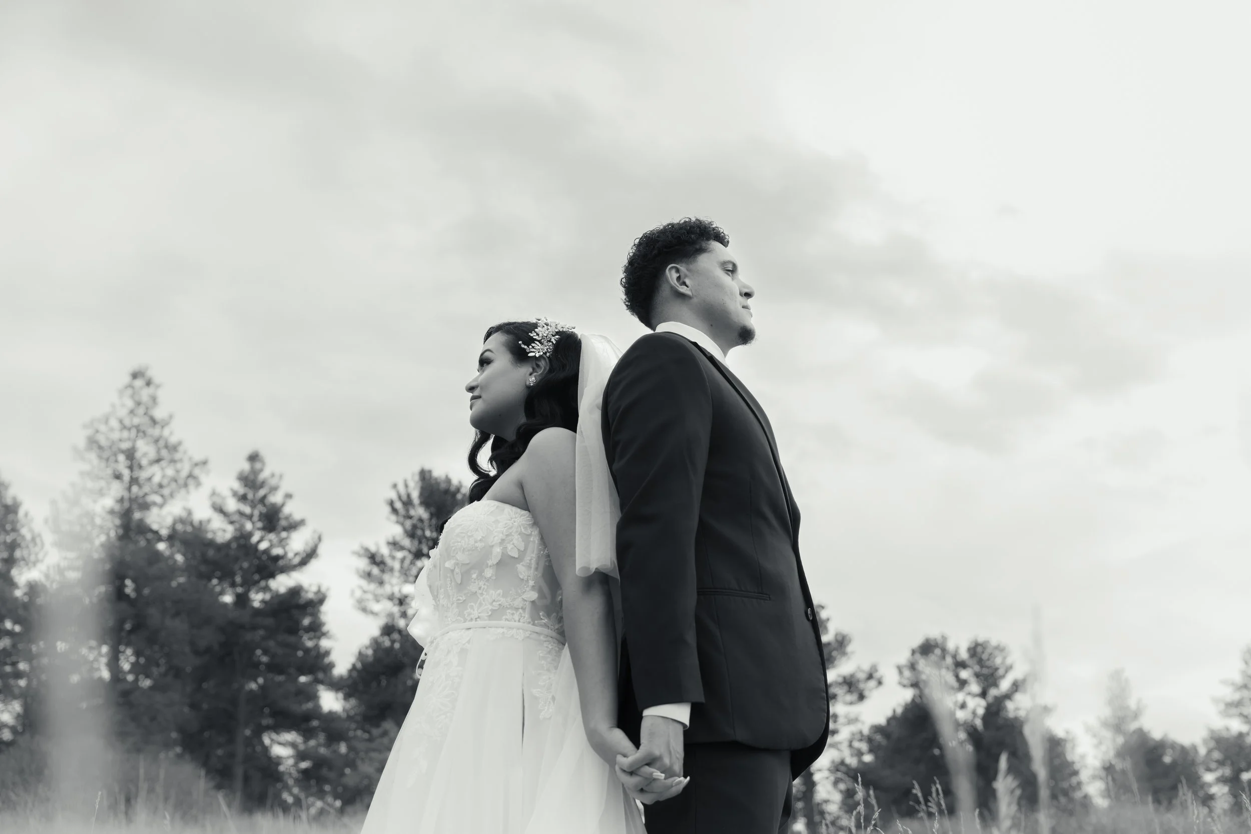A bride and groom standing back-to-back outdoors, holding hands, with trees and cloudy sky in the background, in black and white.