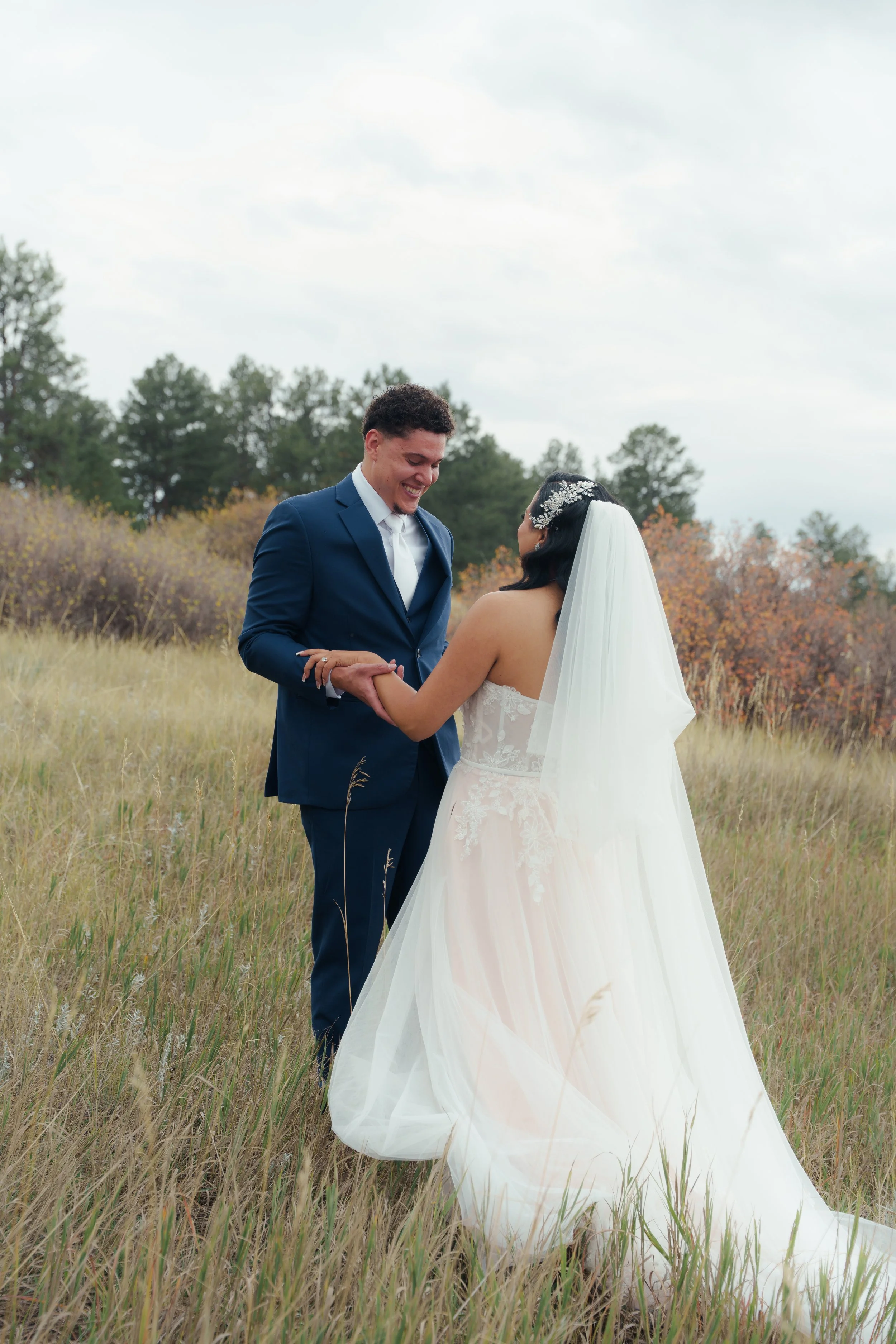 A bride and groom holding hands in a grassy field with trees in the background, during an outdoor wedding.