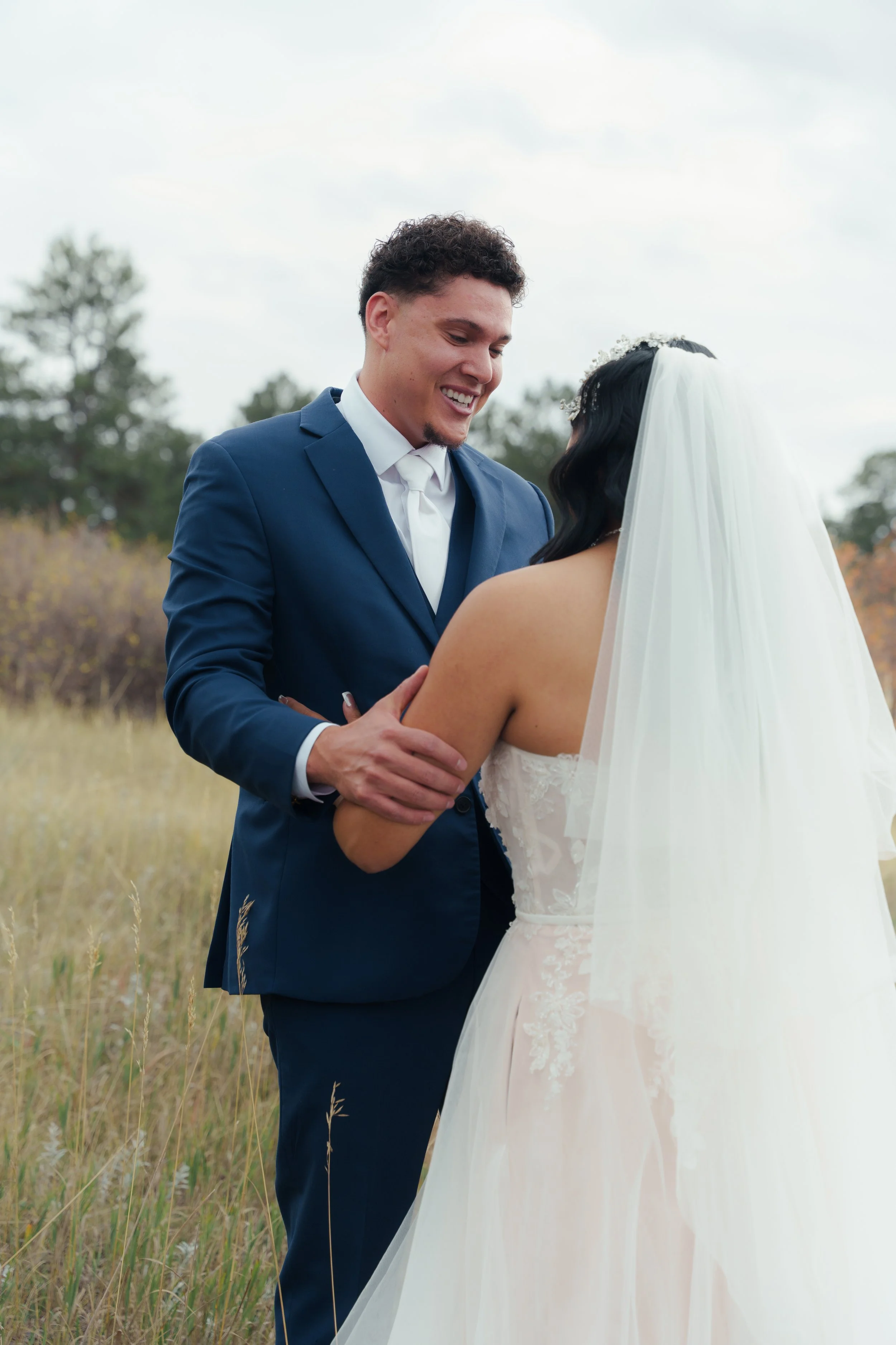 A happy couple on their wedding day outdoors, with the groom in a blue suit and the bride in a white wedding dress and veil, sharing a moment of affection.