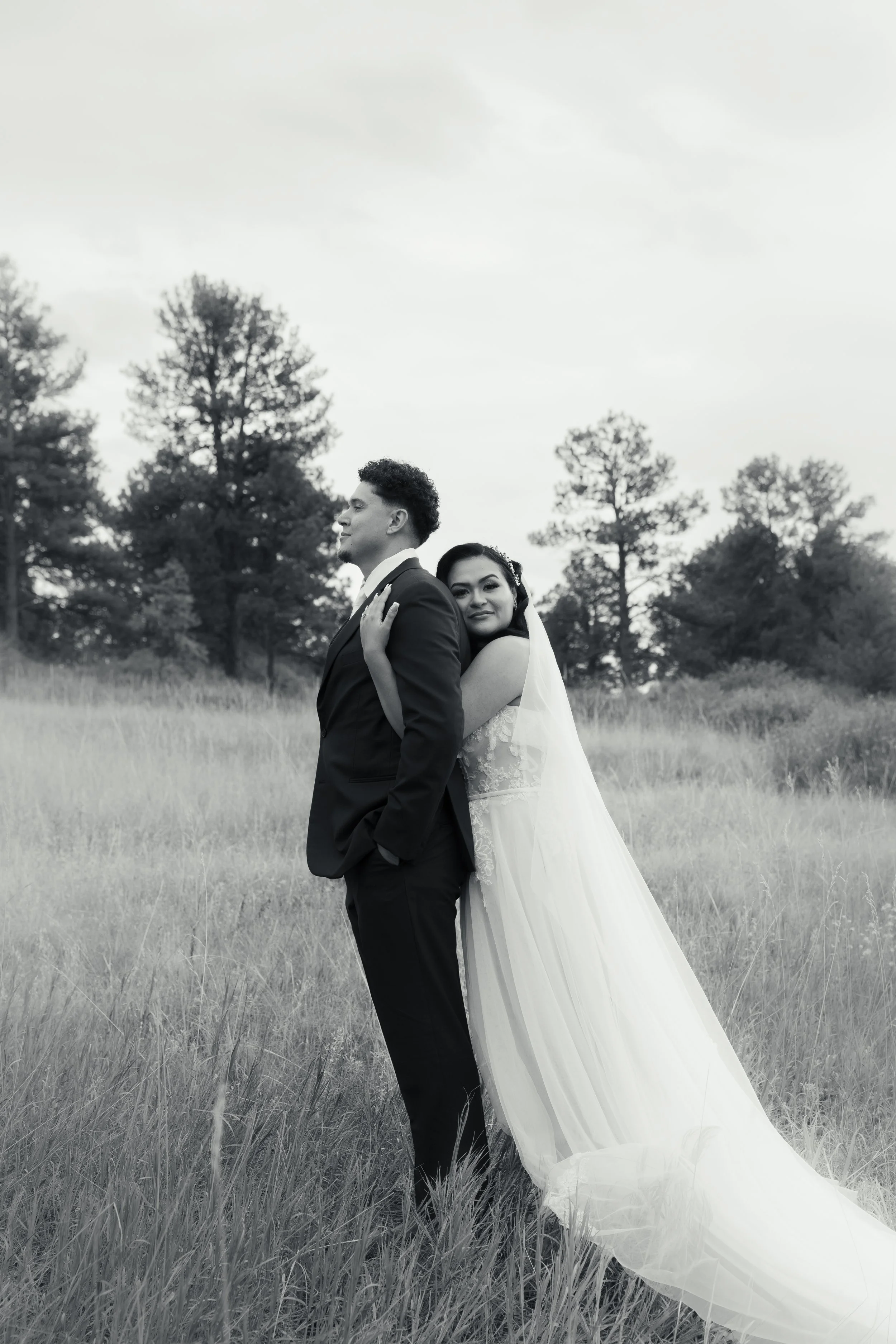 A black and white photo of a bride and groom standing in a field with trees in the background. The bride hugs the groom from behind, resting her head on his back, while he stands with his hands in his pockets and looks to the side.