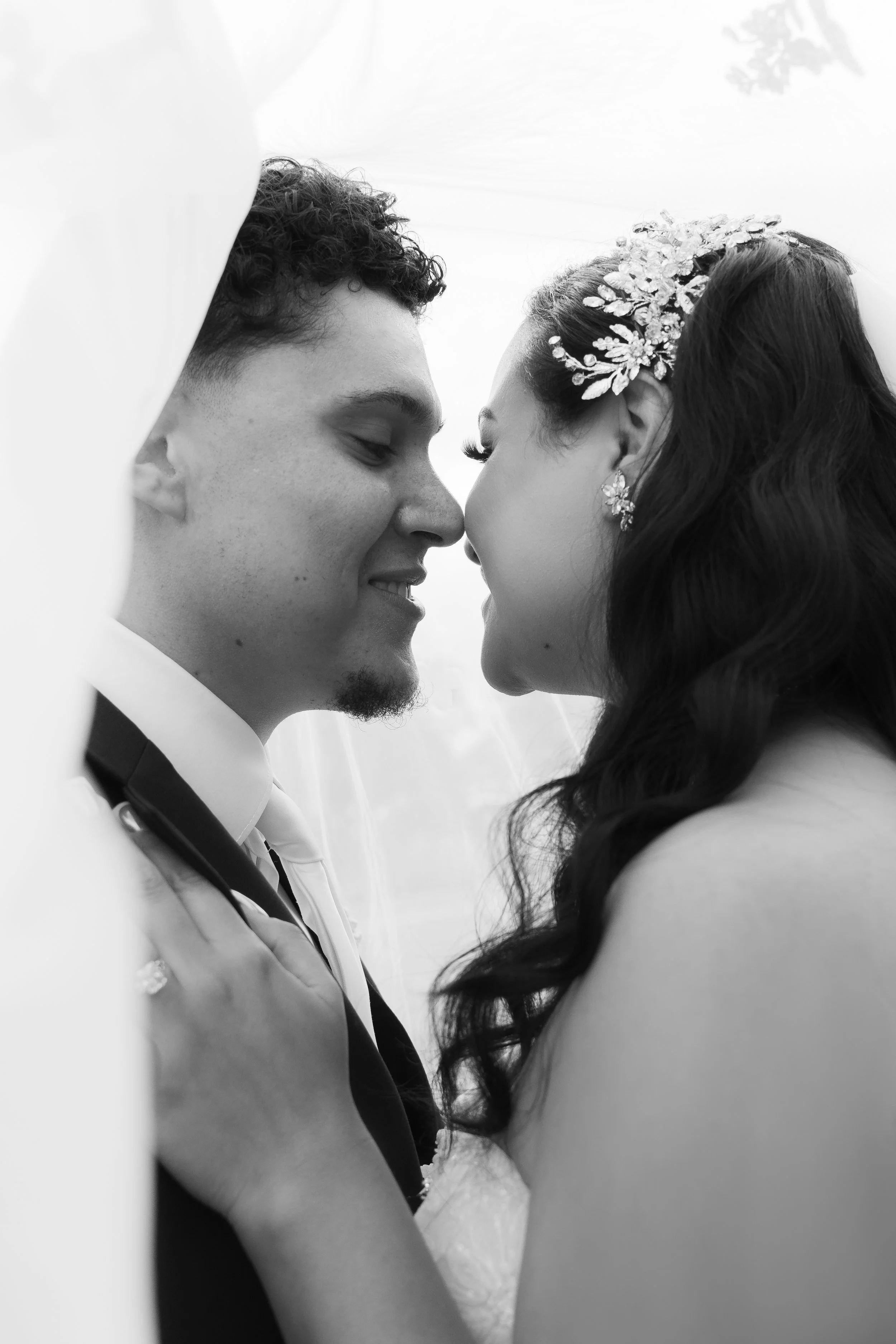Black and white photograph of a couple on their wedding day, close-up with faces touching, smiling, with bridal jewelry and hair accessories.