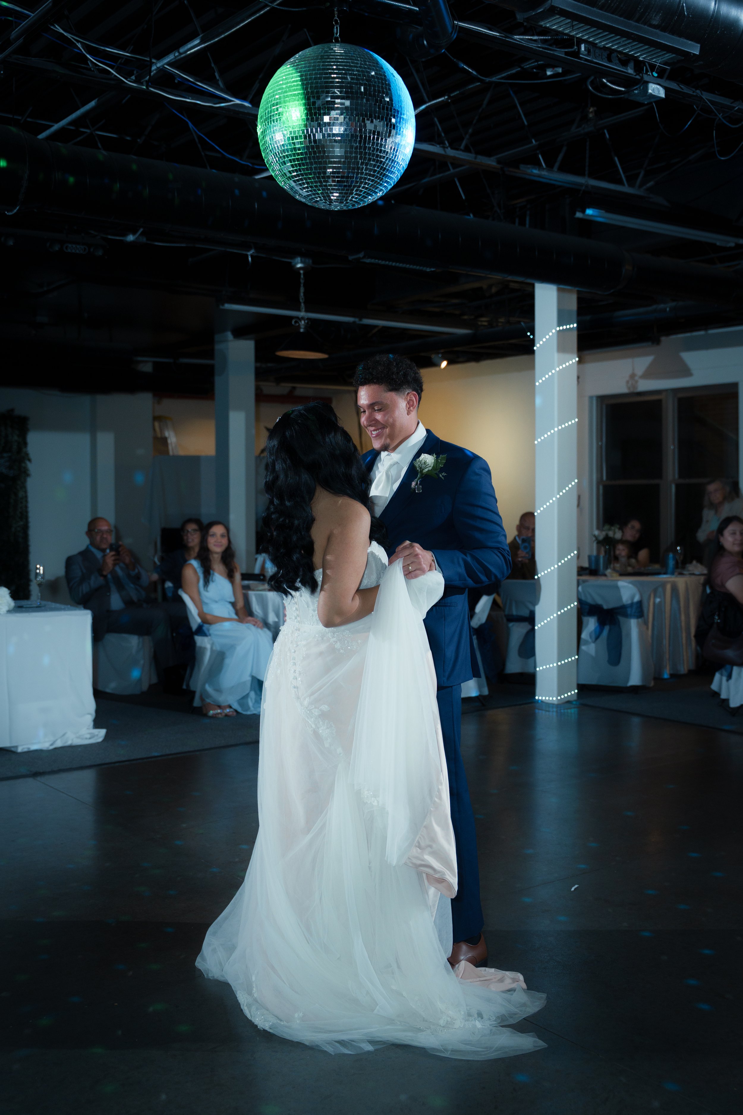 A bride and groom sharing a dance during their wedding reception, with seated guests and a mirror ball overhead.