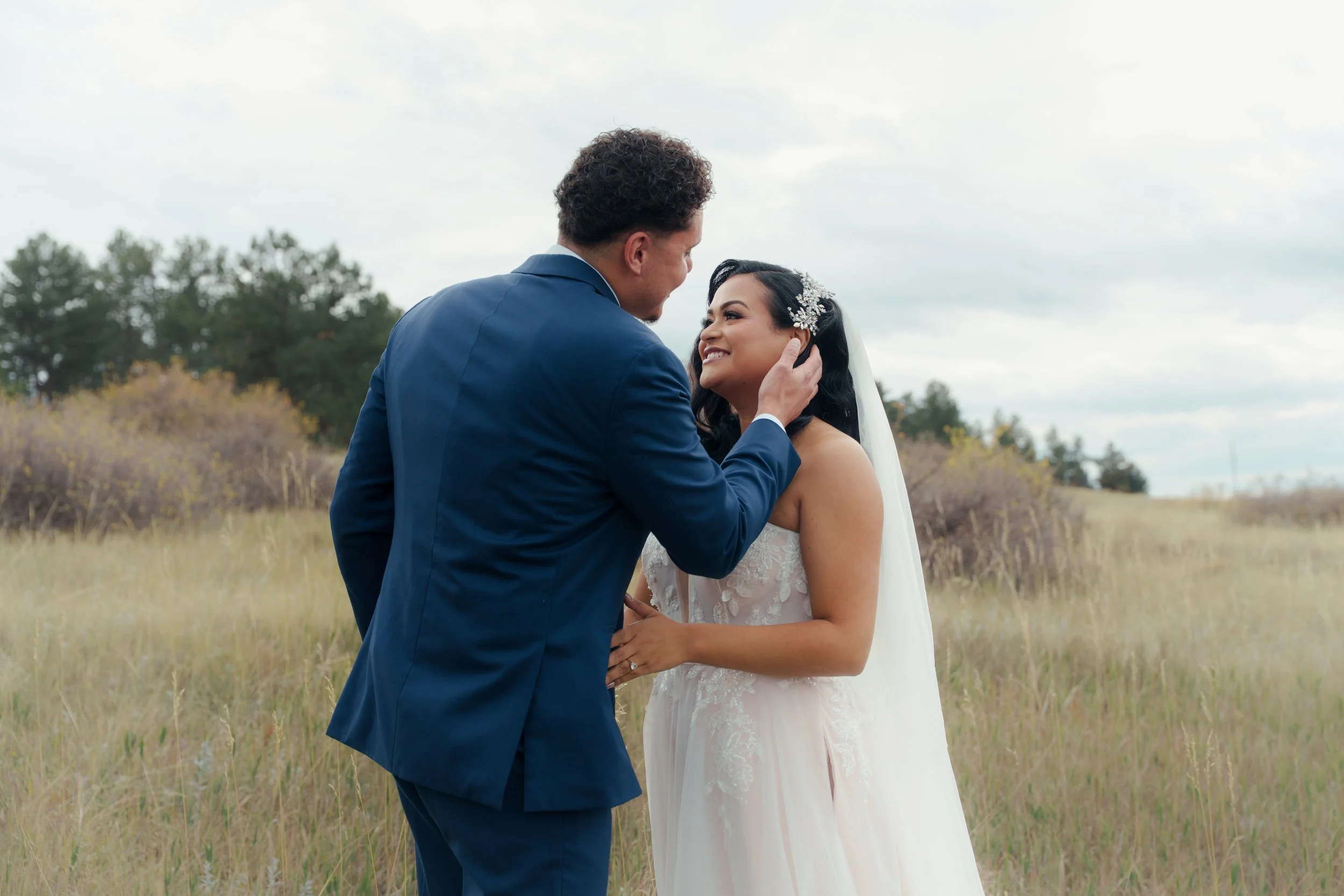 A bride and groom standing in a field, looking at each other and smiling, during their wedding.