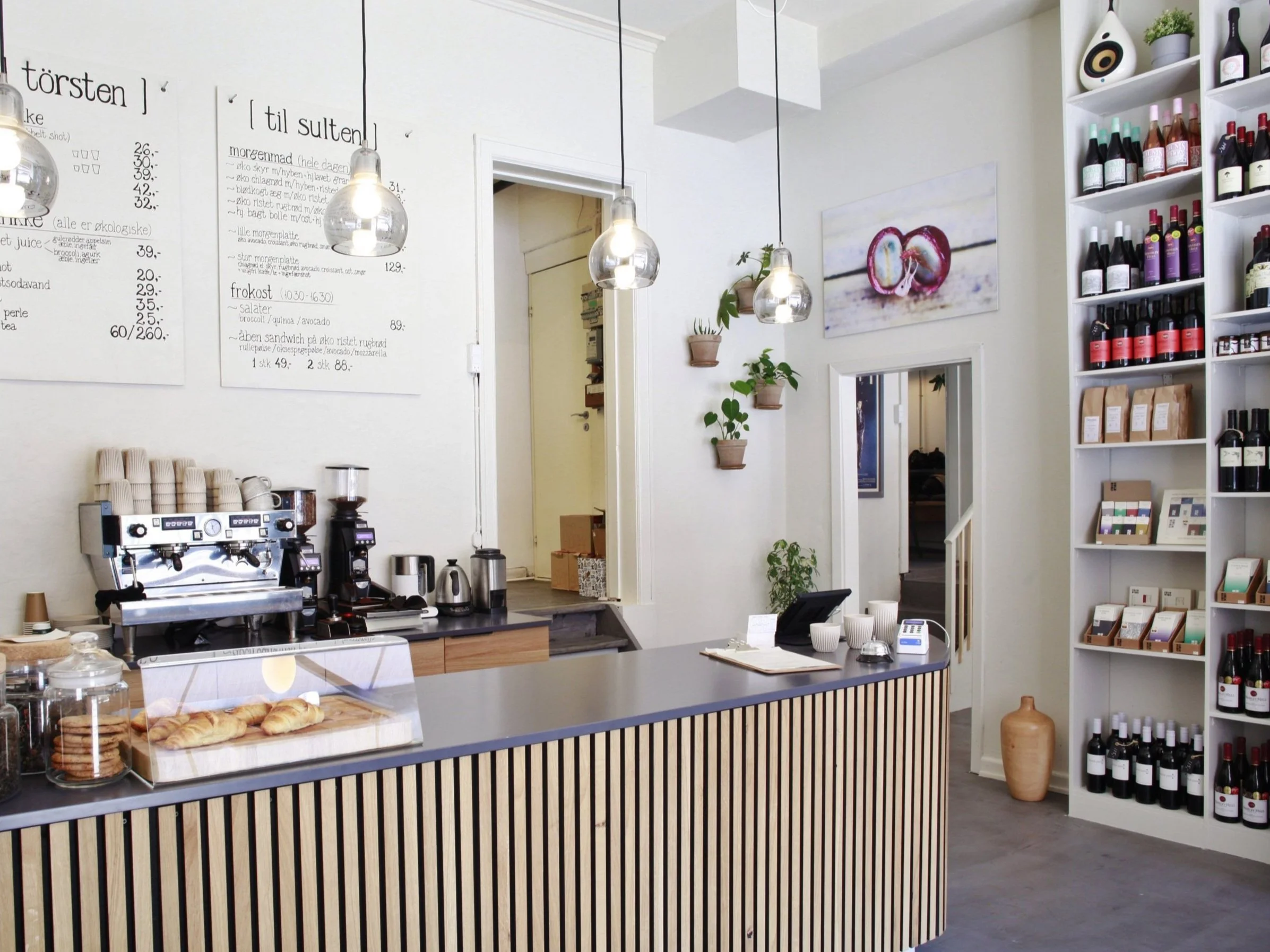 Interior of a modern cafe with a counter featuring an espresso machine, cups, pastries, and a menu on the wall. Shelves with wine bottles and packaged goods are on the side. Hanging light fixtures and potted plants add decorative touches.