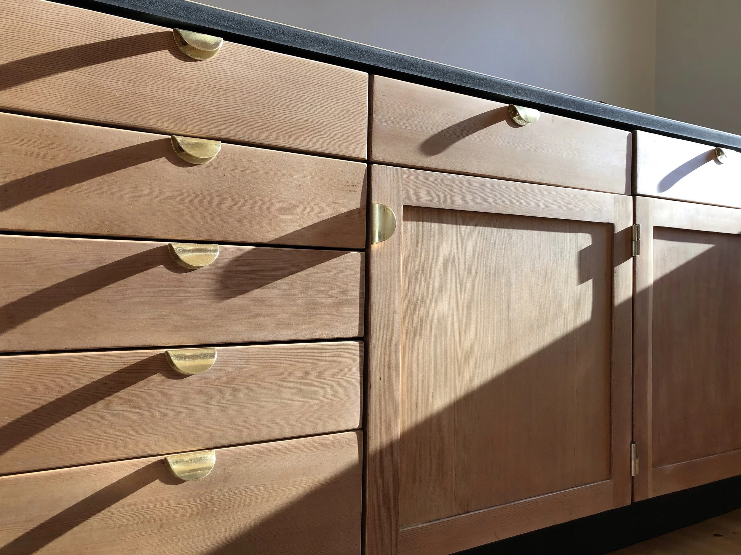 Wooden kitchen cabinets with brass handles and a dark countertop, illuminated by sunlight, creating shadows on the surface.