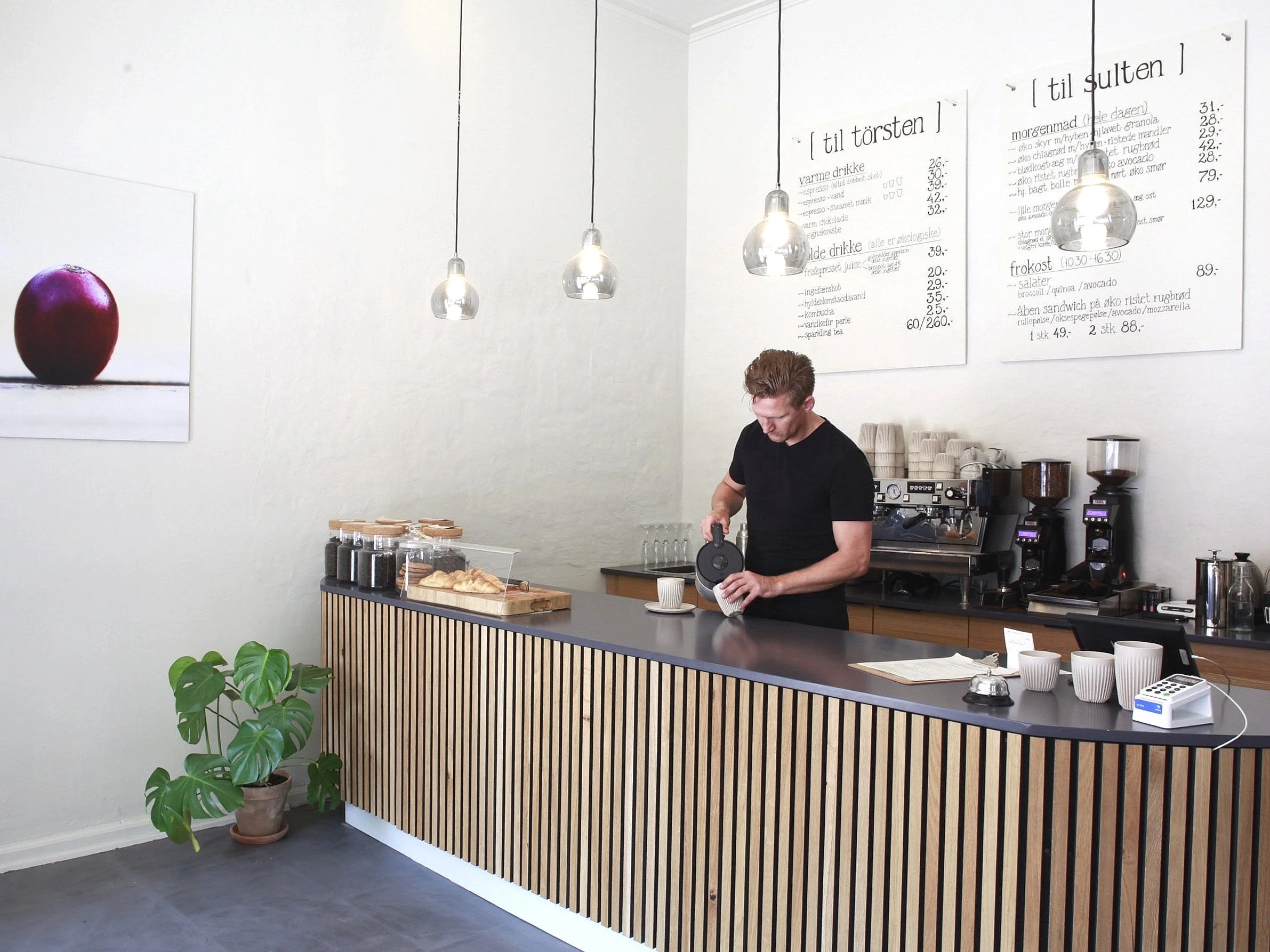 Barista pouring coffee in a minimalist cafe with a wooden counter, coffee jars, and a menu on the wall.