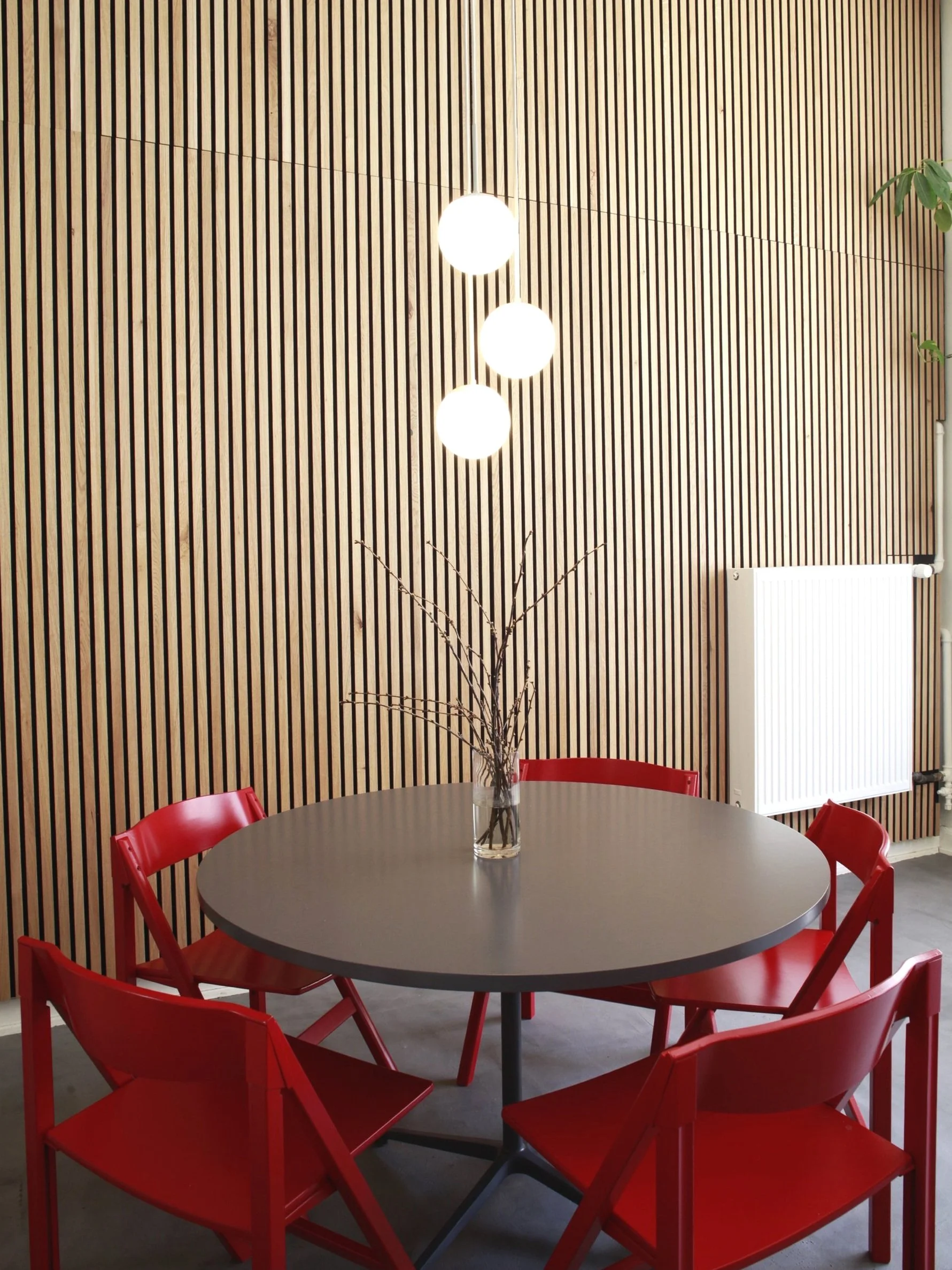 Modern dining area with a round gray table surrounded by four red chairs. A glass vase with branches is on the table. Background features a wooden slatted wall and three spherical hanging lights.