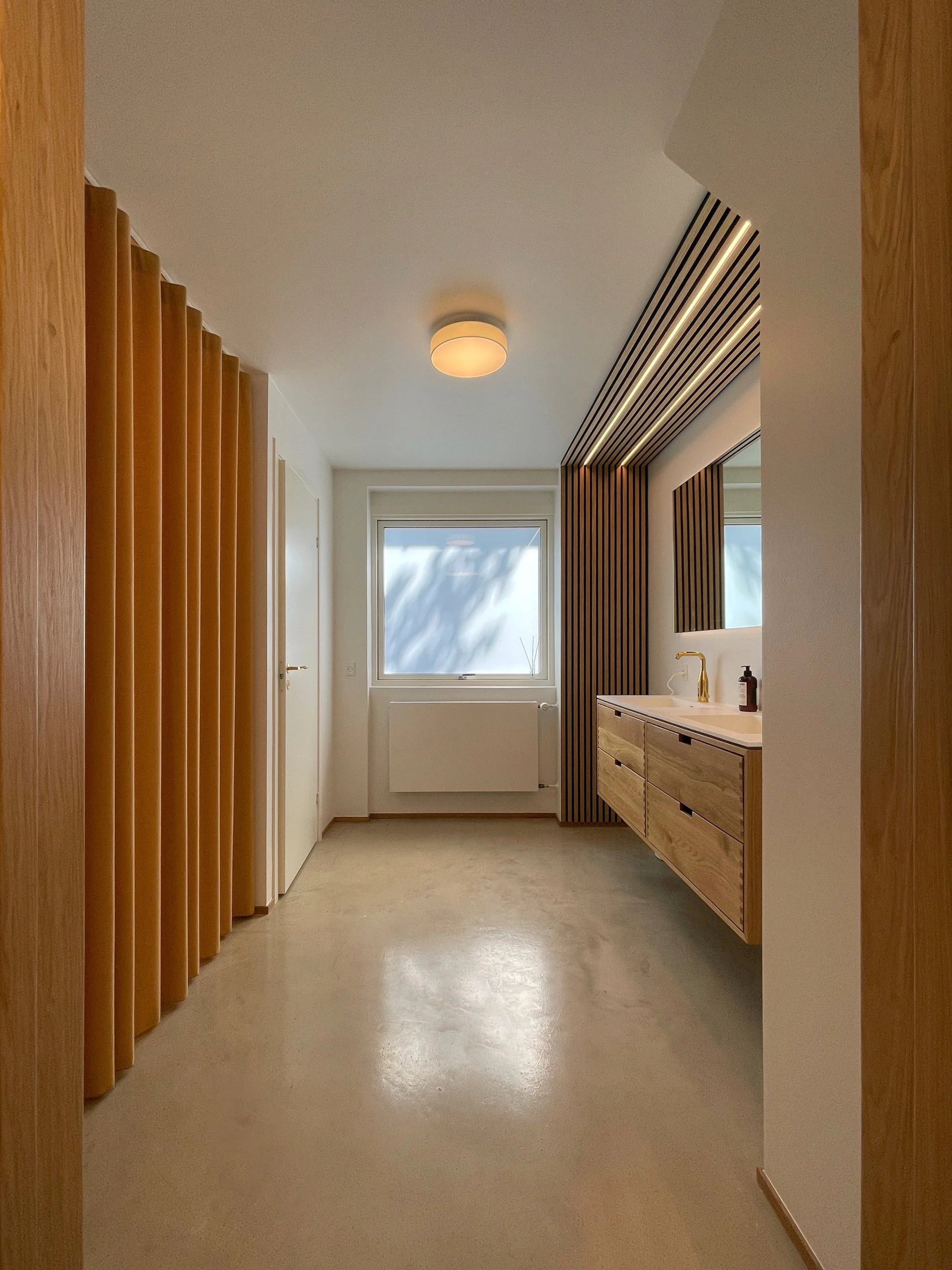 Minimalist bathroom with wooden accents featuring a floating vanity, a large mirror, and a window with natural light. The ceiling light and vertical slats enhance the clean, modern design.
