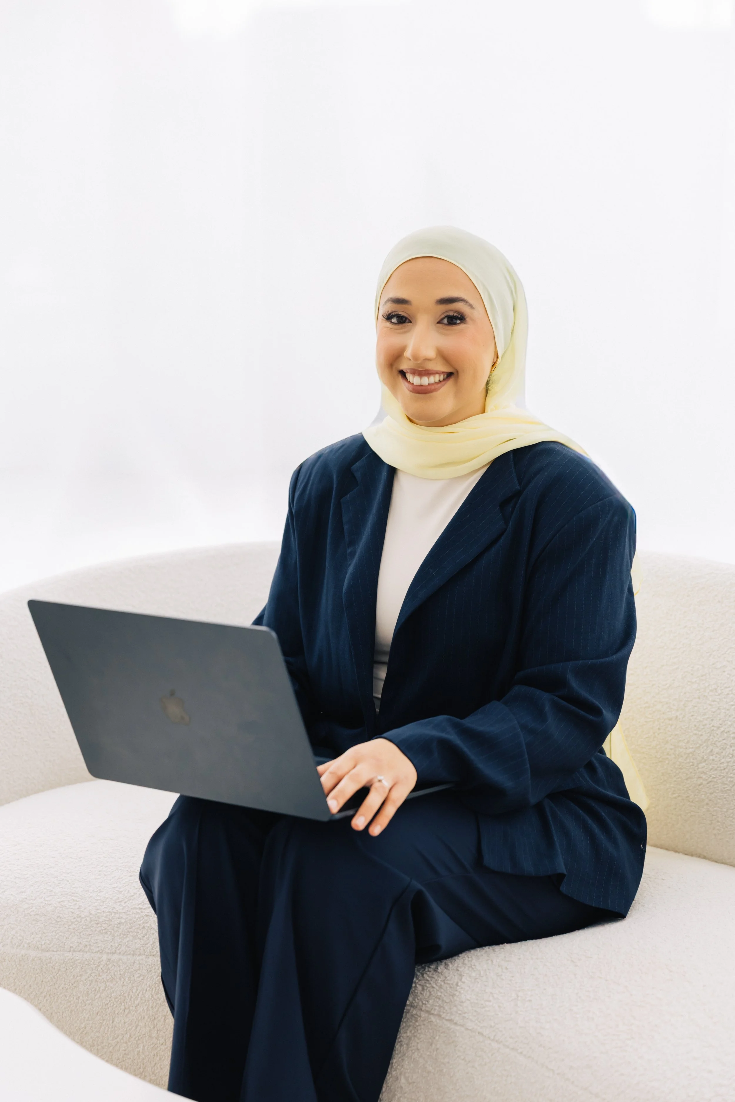 A smiling woman wearing a light yellow headscarf and navy blue business suit sitting on a cream-colored couch with a laptop on her lap.