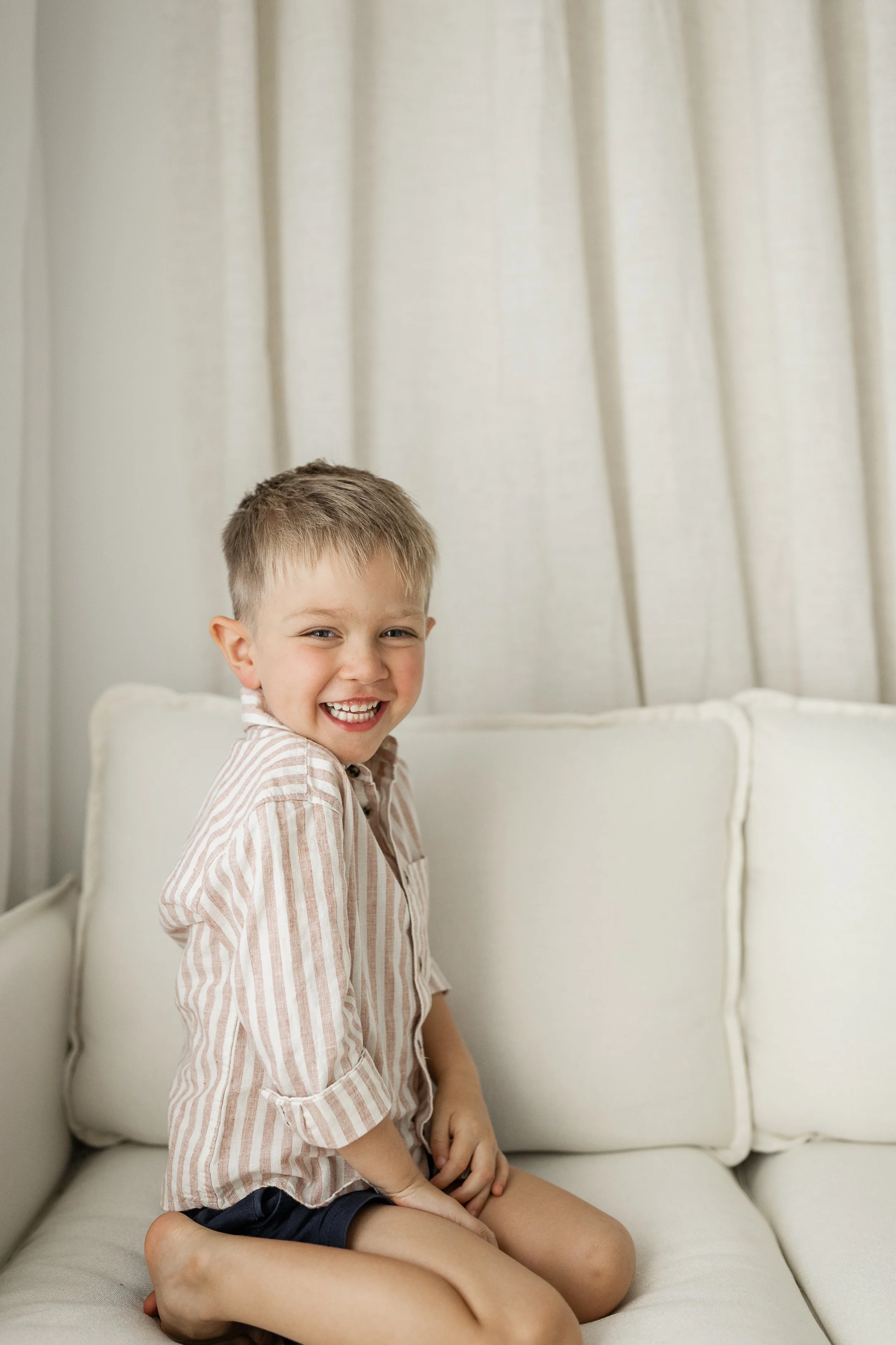 Smiling young child seated on a white sofa with a light-colored striped shirt and shorts.