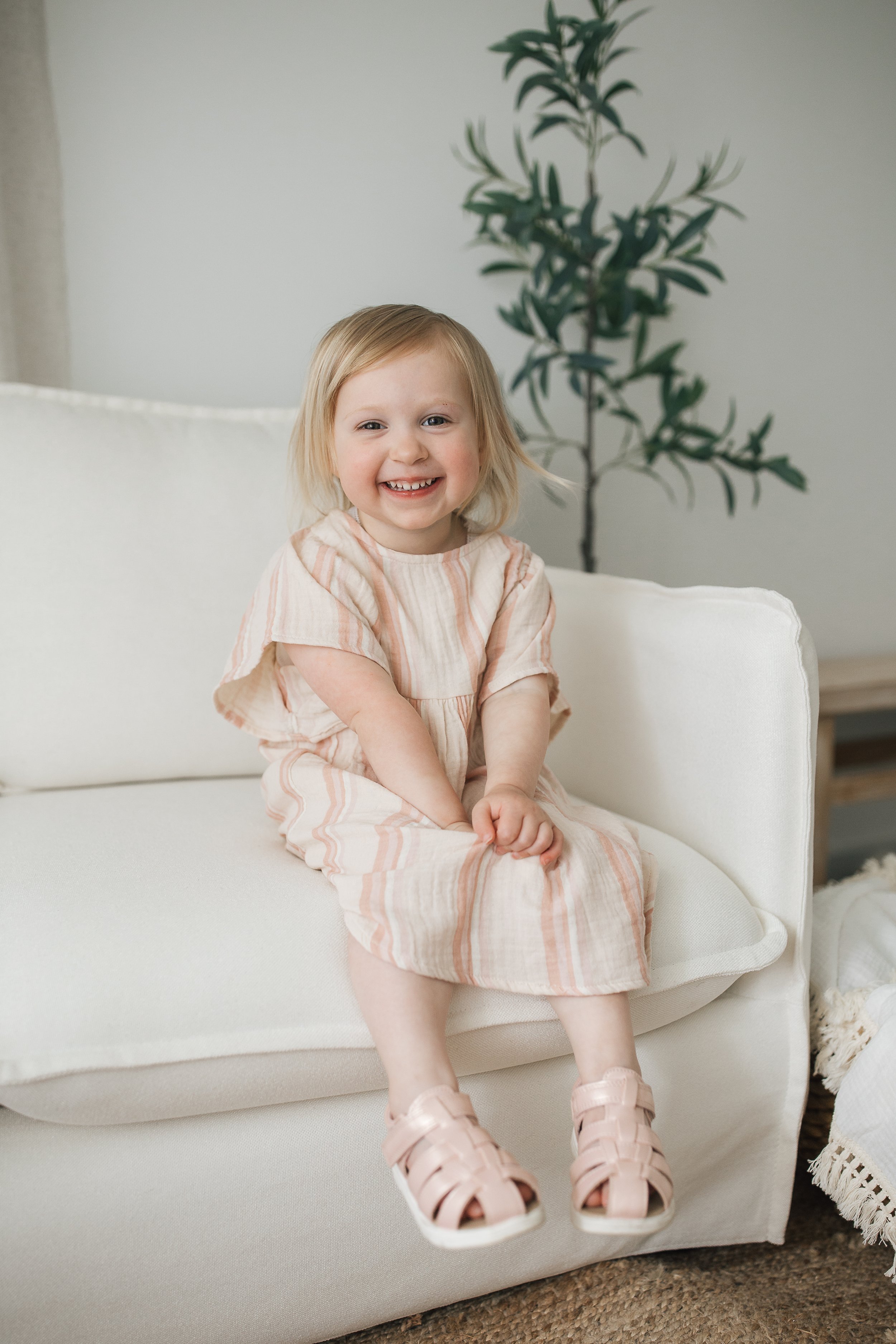 Young child sitting on a white couch, wearing a striped dress and pink sandals, smiling with an indoor plant in the background.