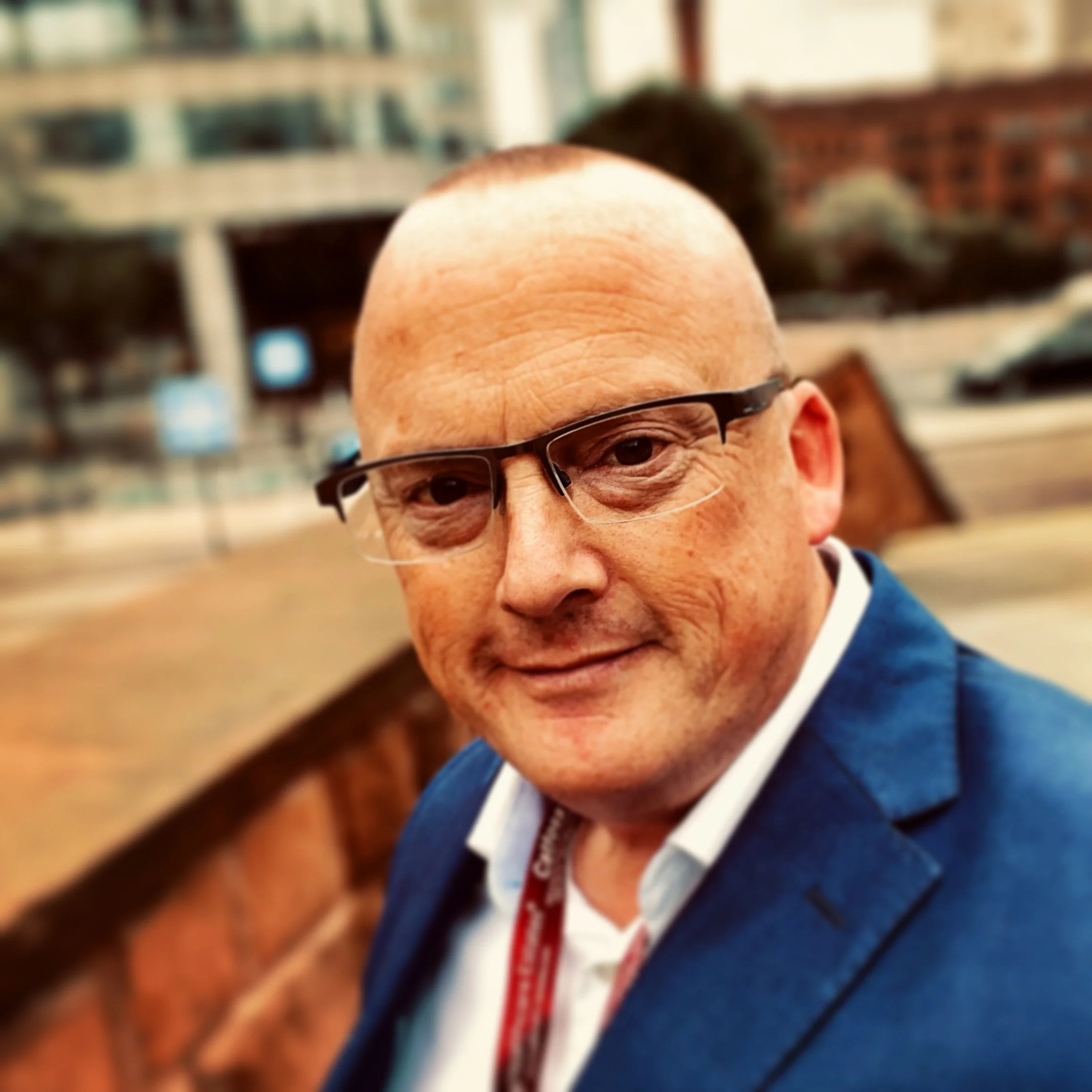 A middle-aged man with glasses, a bald head, and a smile, wearing a blue suit jacket and white shirt, sitting at a wooden table in a busy conference or event setting.