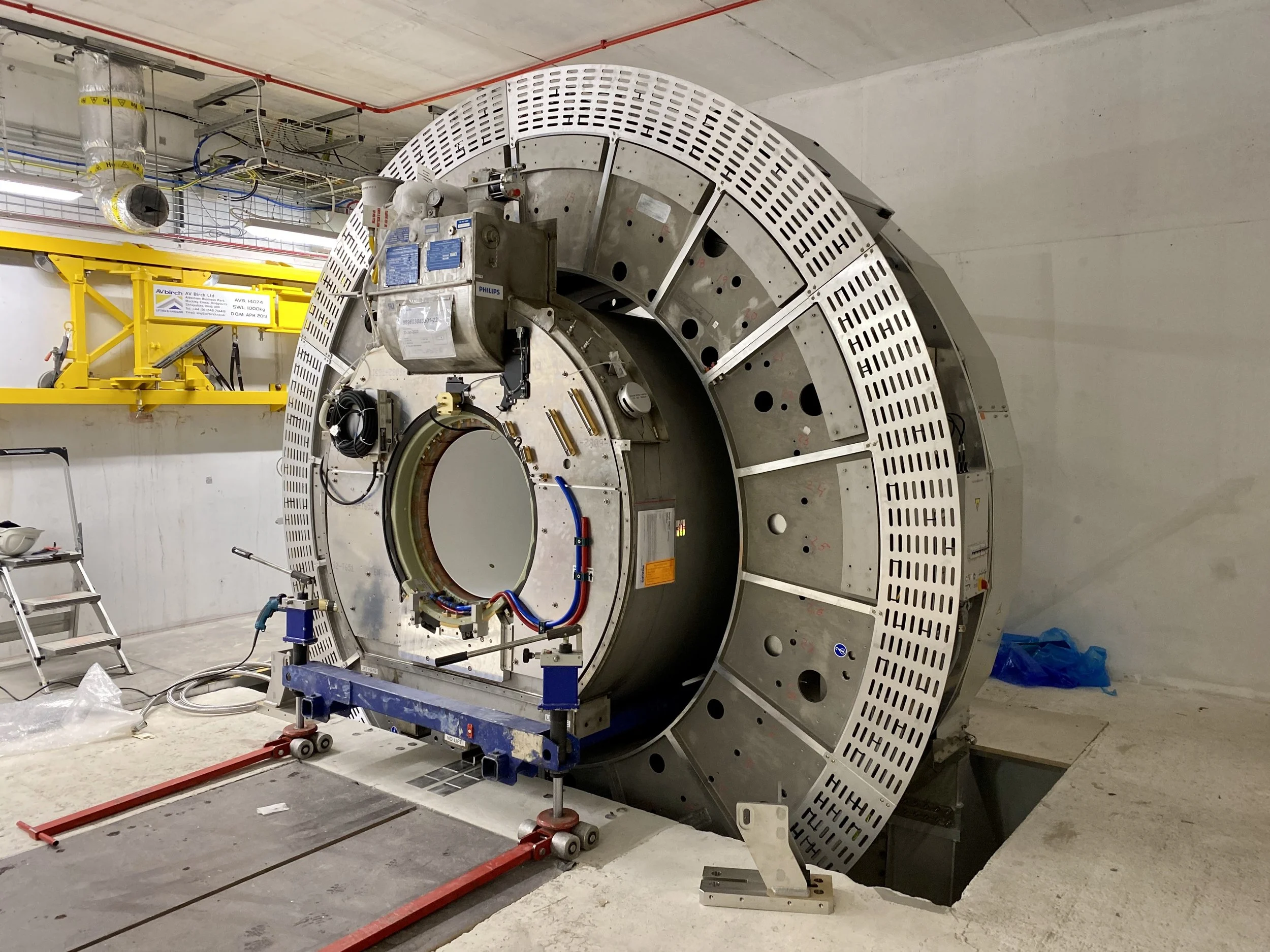 Large circular piece of industrial equipment with various wiring, mounting brackets, and small components, situated in a construction or laboratory setting with tools and a yellow crane in the background.