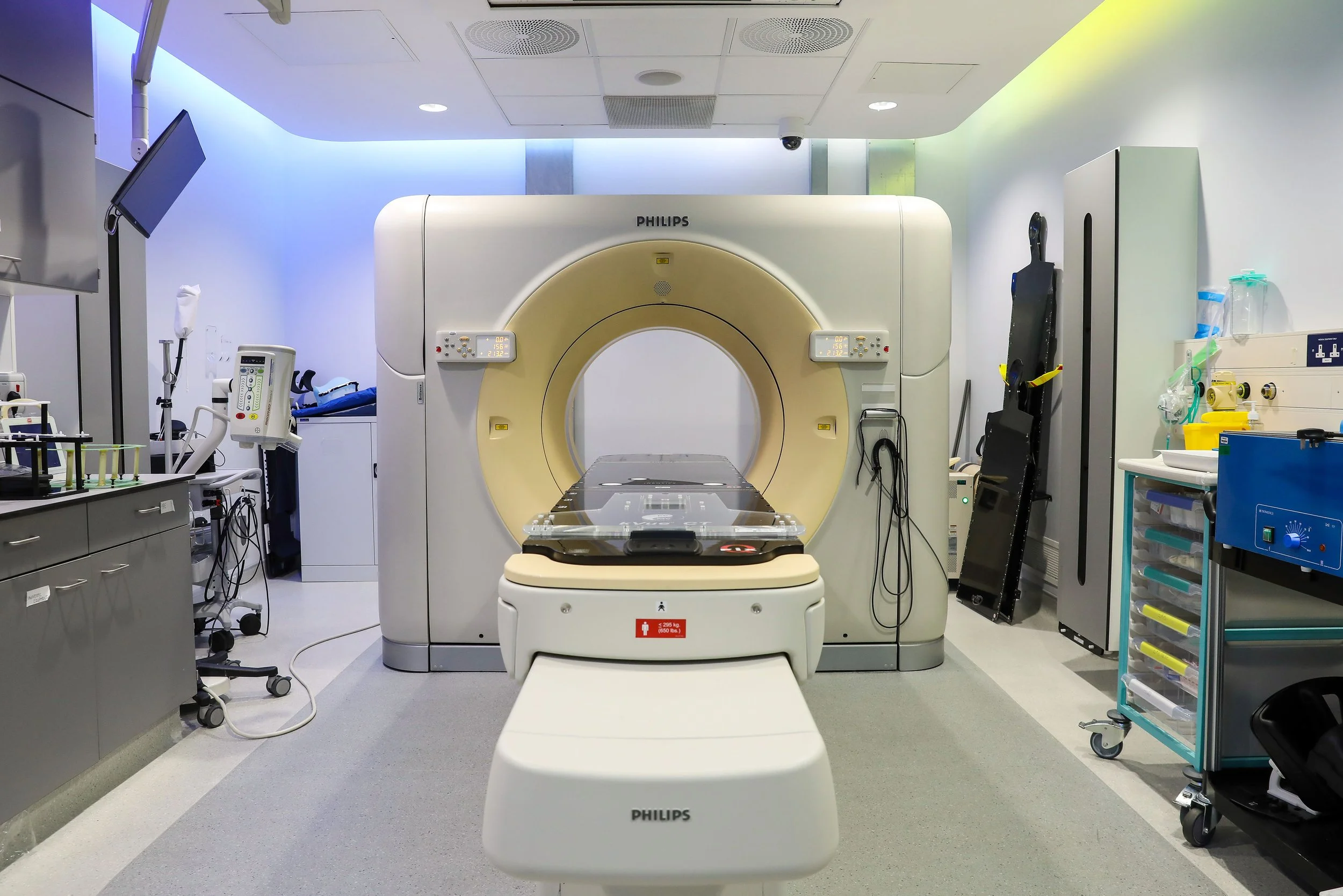 Medical imaging room with a large Philips CT scanner, a padded table in front, and various medical equipment and supplies on counters and carts.