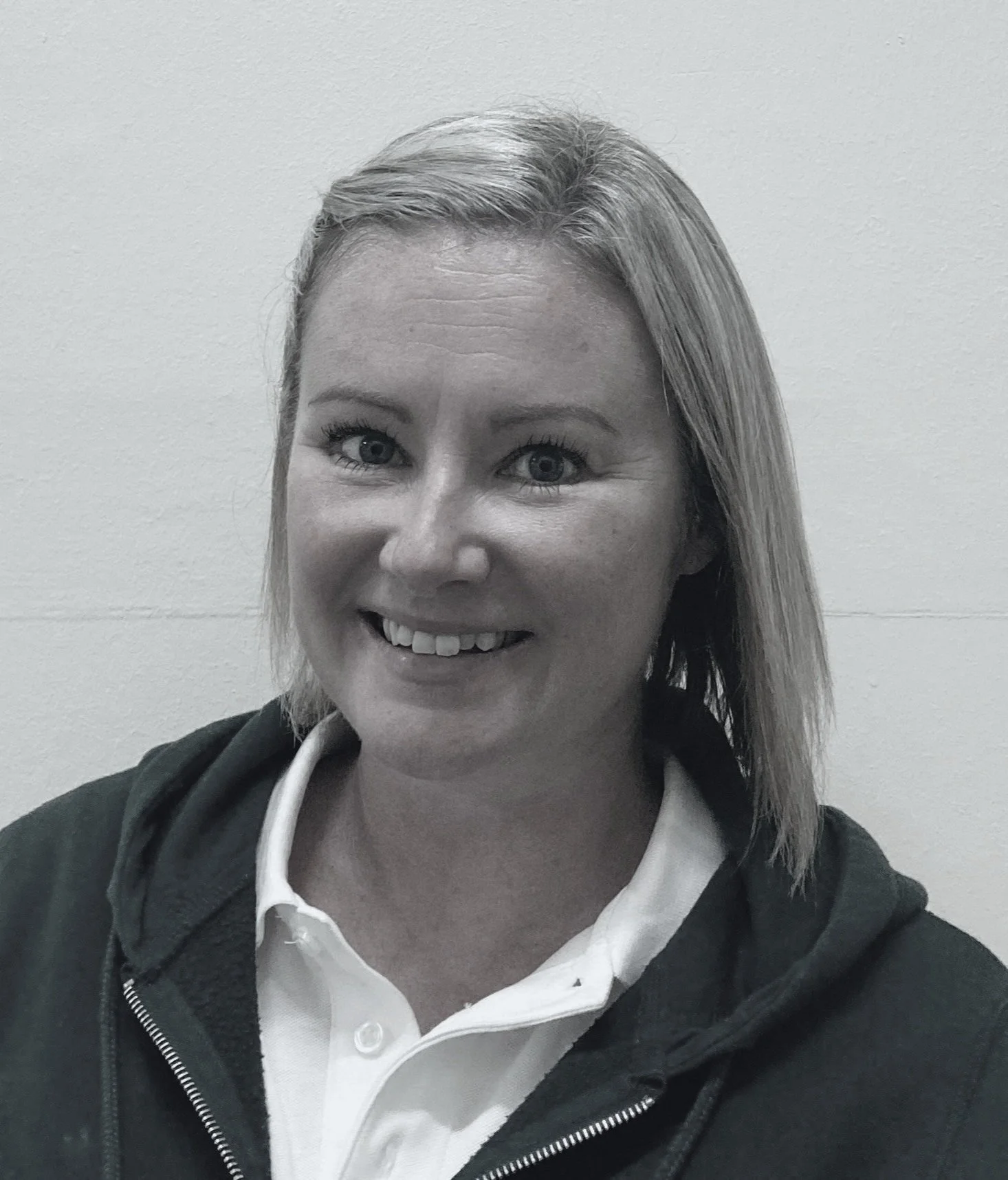 A woman smiling, wearing a white collared shirt and a dark zip-up hoodie, with short blonde hair, against a plain light-colored wall.