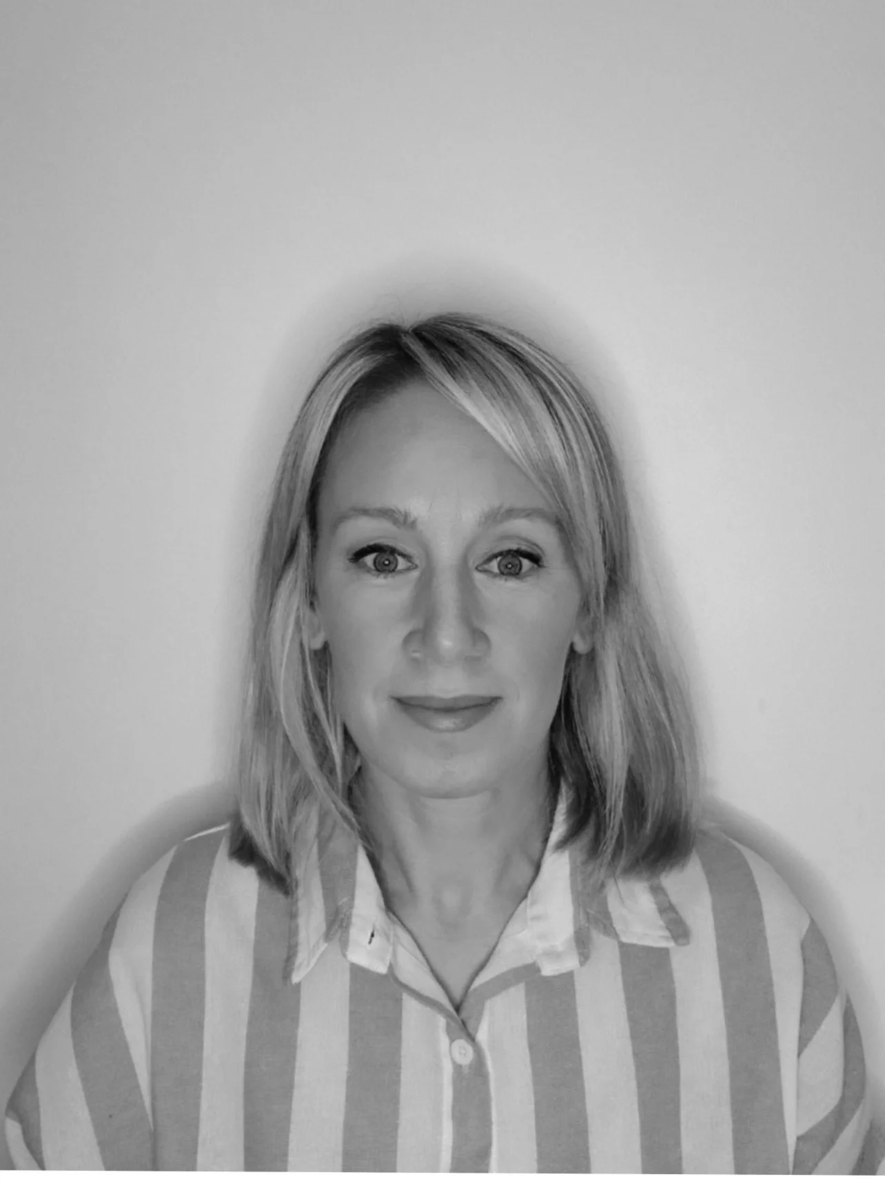 Black and white portrait of a woman with shoulder-length hair, wearing a striped collared shirt, standing against a plain background.