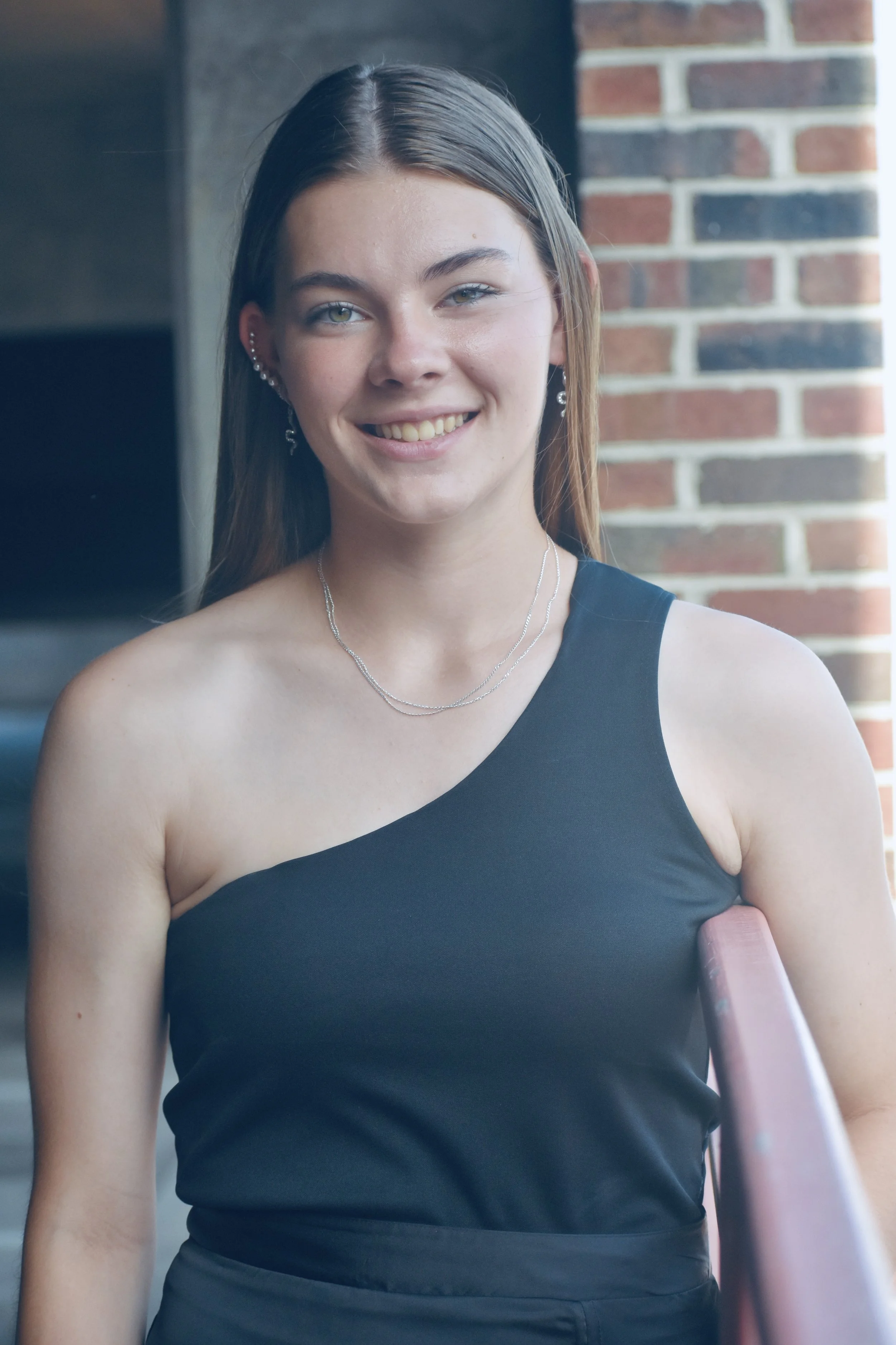 A young woman with long brown hair, wearing a black sleeveless top, standing outdoors against a brick wall, smiling, with a necklace and earrings.