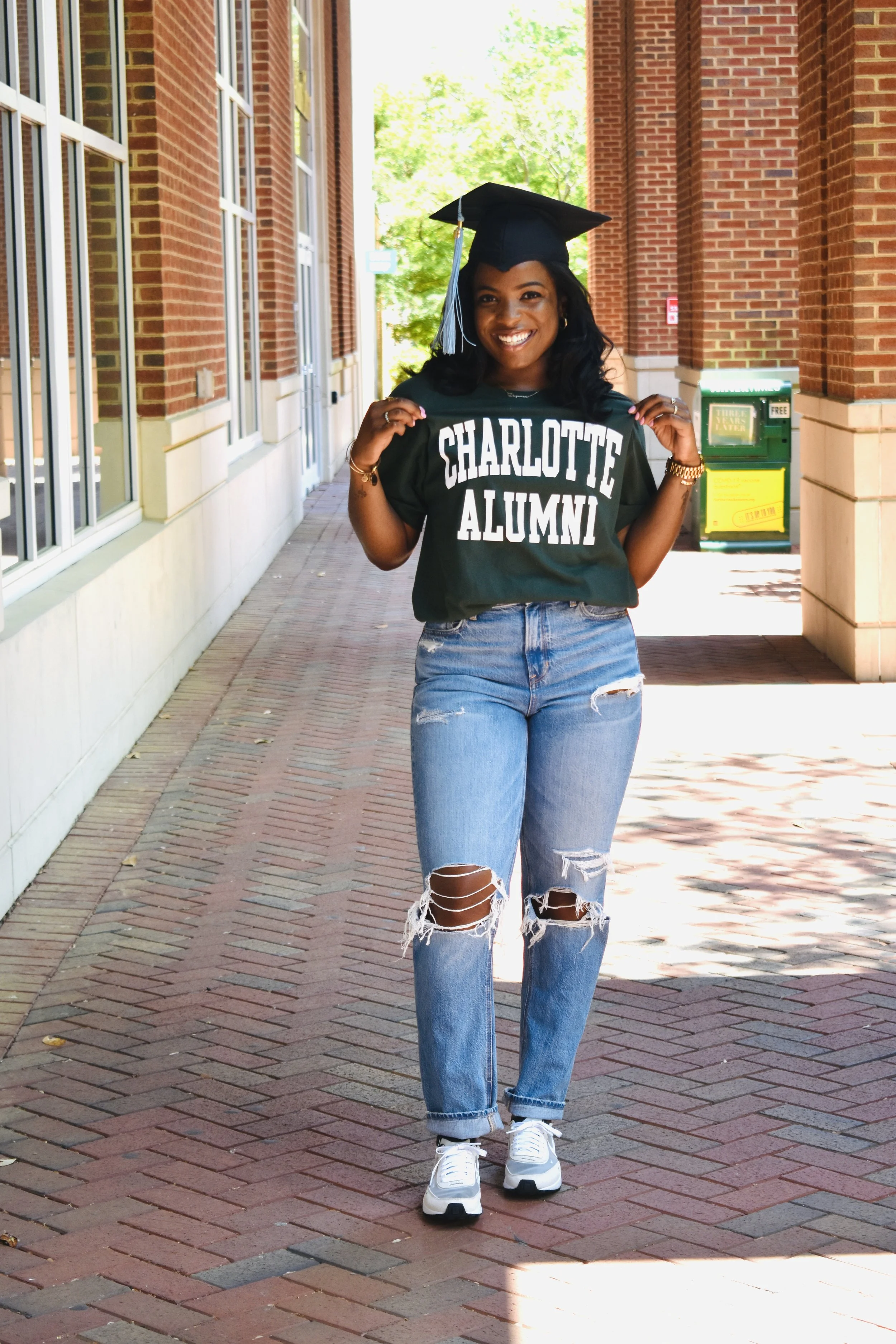 A young woman wearing a black graduation cap and a green t-shirt that says 'Charlotte Alumni' is smiling and standing on a brick sidewalk outside a brick building with large windows.