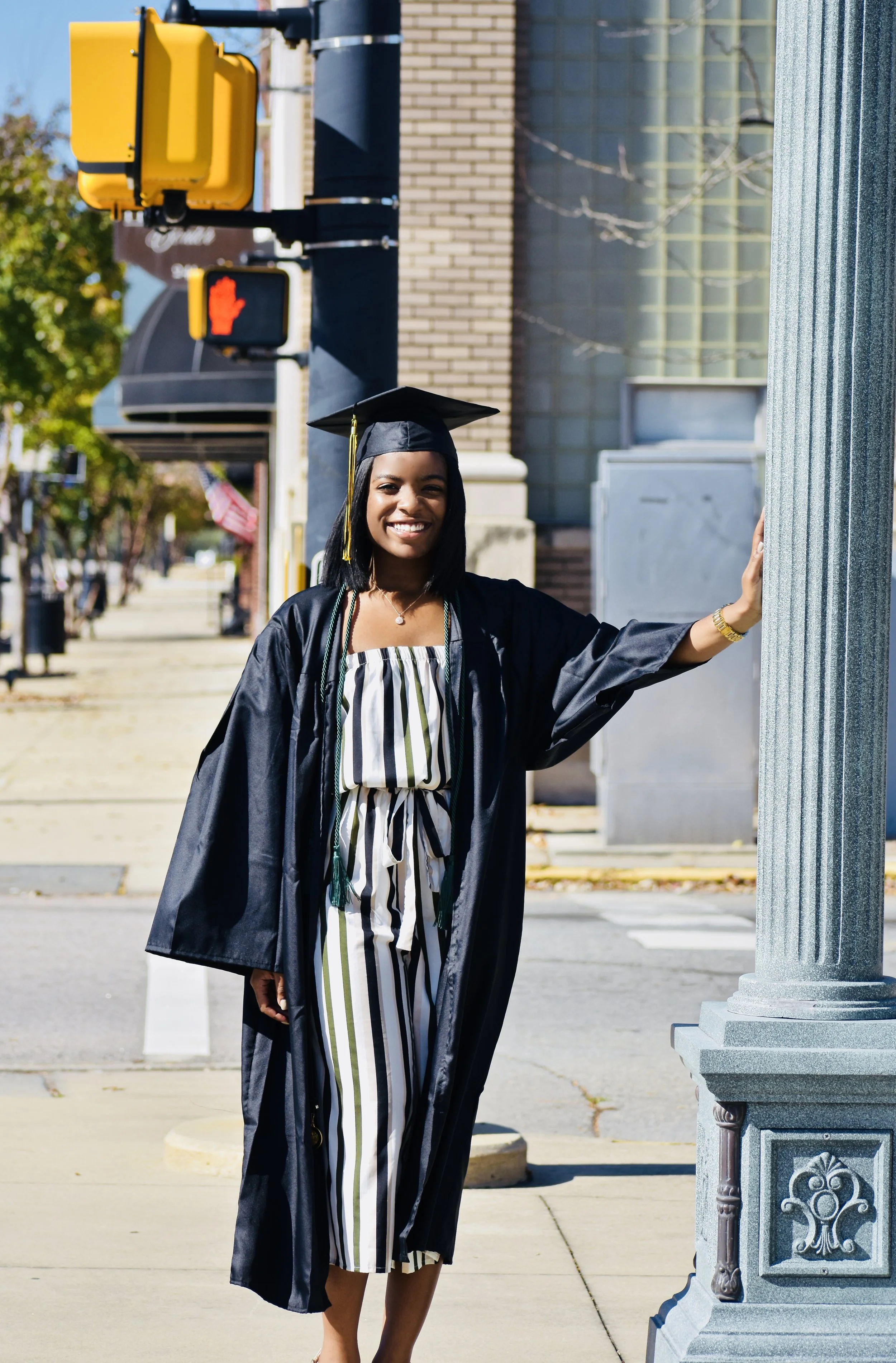 A young woman in a graduation cap and gown smiling while standing outside on a sunny day, leaning on a grey pillar near a street with traffic lights and buildings in the background.