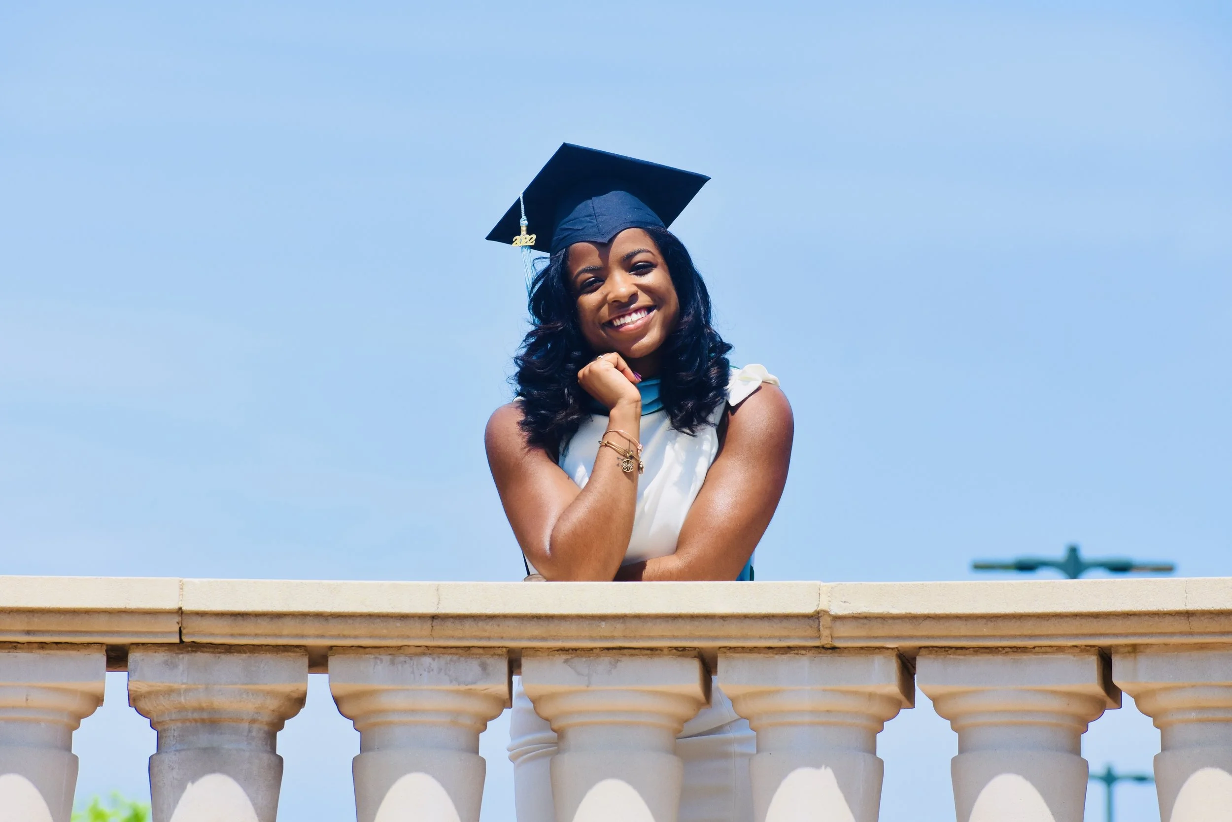 A young woman in a graduation cap and dress leaning on a beige balcony rail, smiling against a clear blue sky.