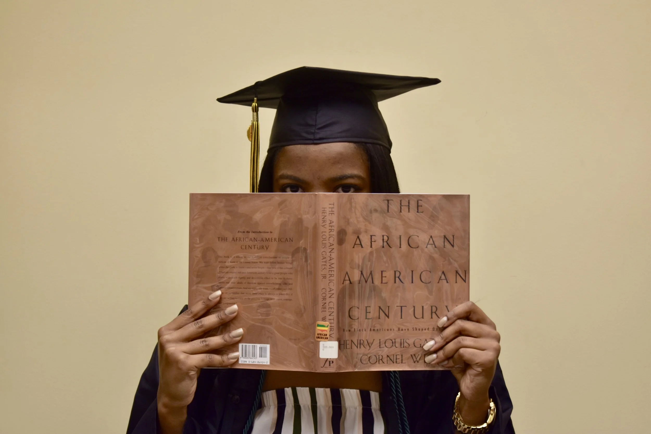 A young woman in graduation attire wearing a black cap and gown, holding a book titled 'The African American Century' in front of her face, with only her eyes visible, standing against a plain beige wall.