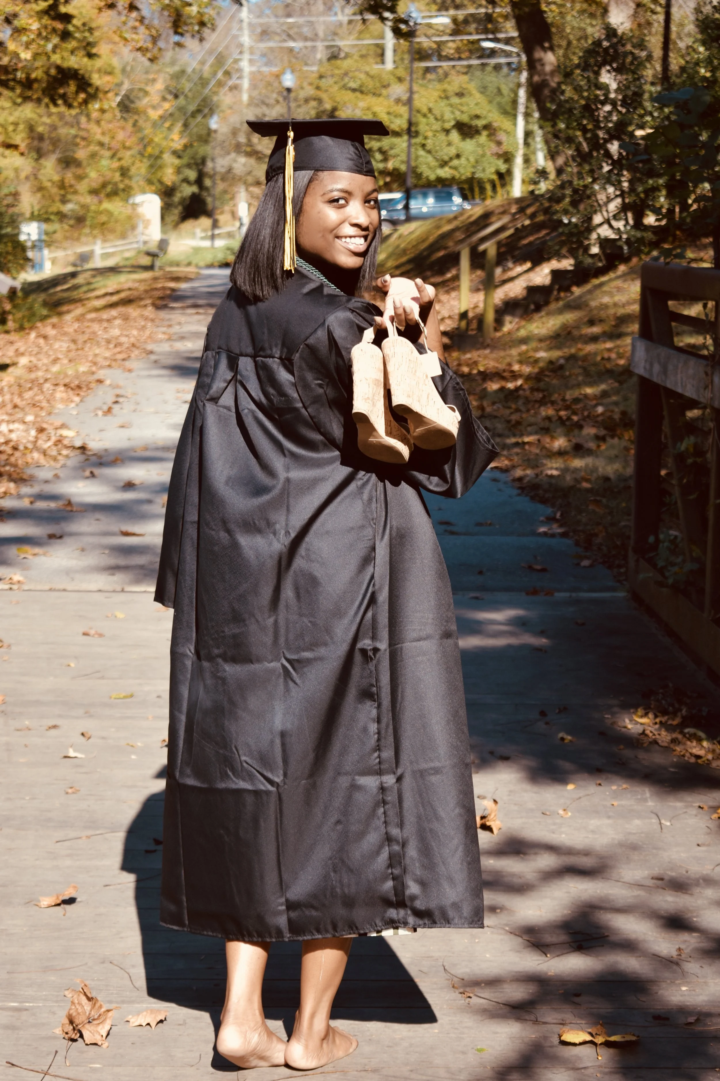 A young woman in a black graduation gown and cap holding a pair of shoes, walking barefoot on a leaf-covered path outdoors during fall.