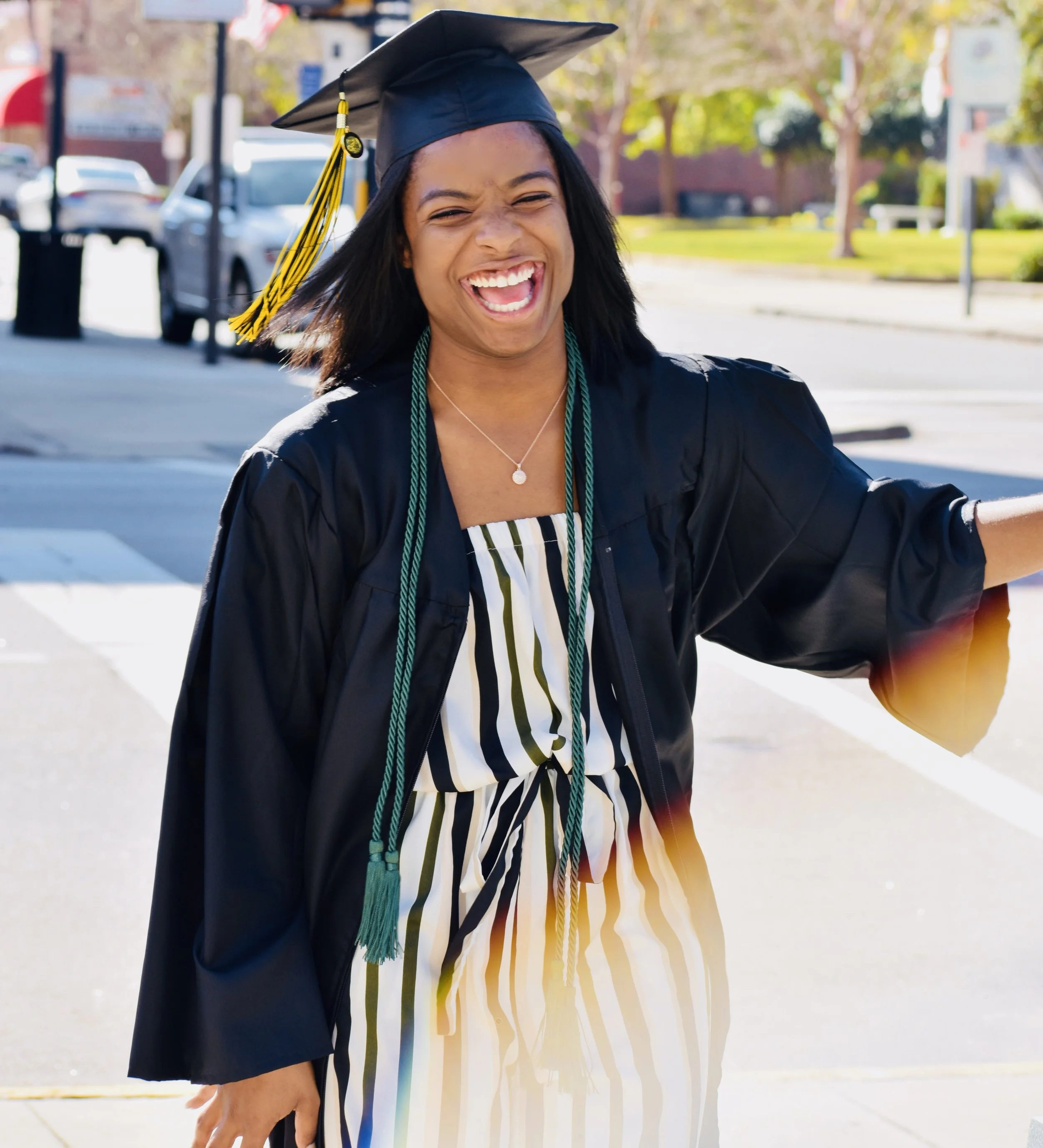 Young woman in graduation cap and gown celebrating outdoors on a sunny day.