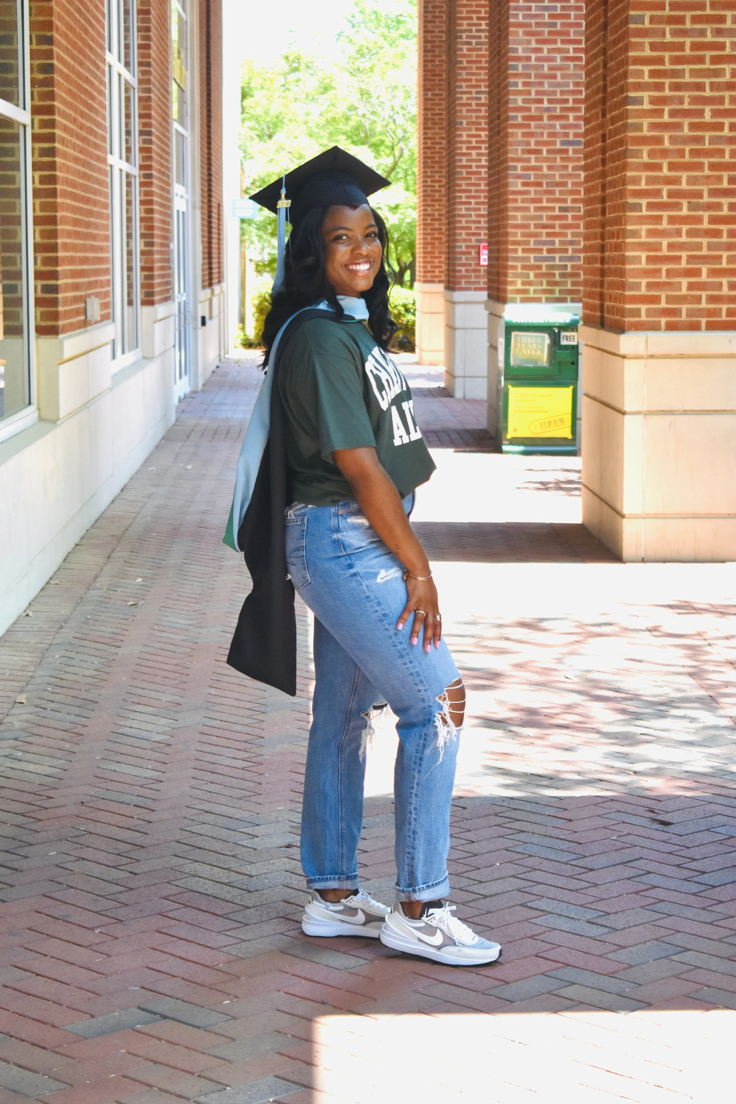 A young woman in a graduation cap and gown standing on a brick sidewalk outside a university building, smiling at the camera.