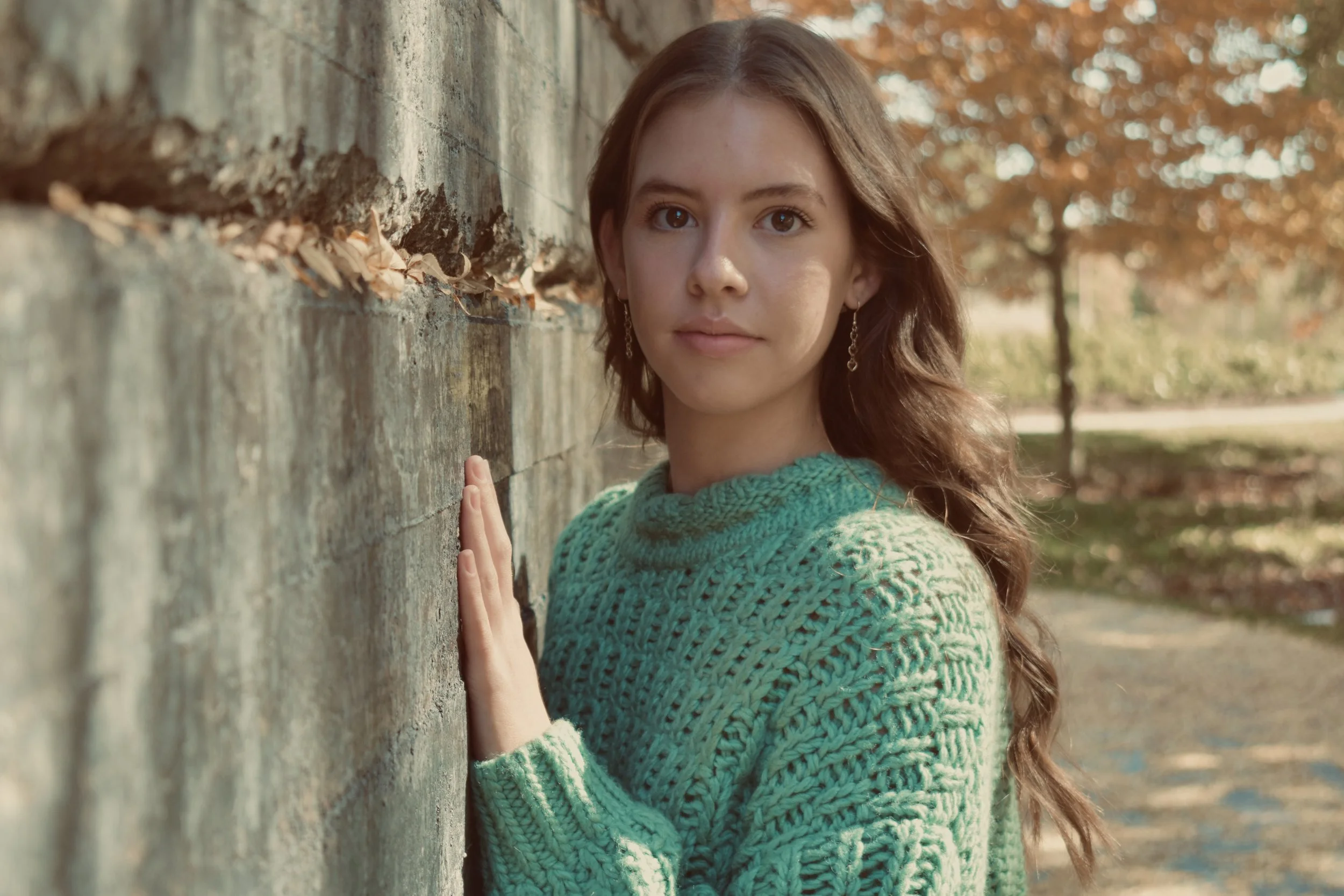 A young woman with long wavy hair, wearing a green knitted sweater and earrings, standing against a stone wall outdoors during fall, with trees and fallen leaves in the background.
