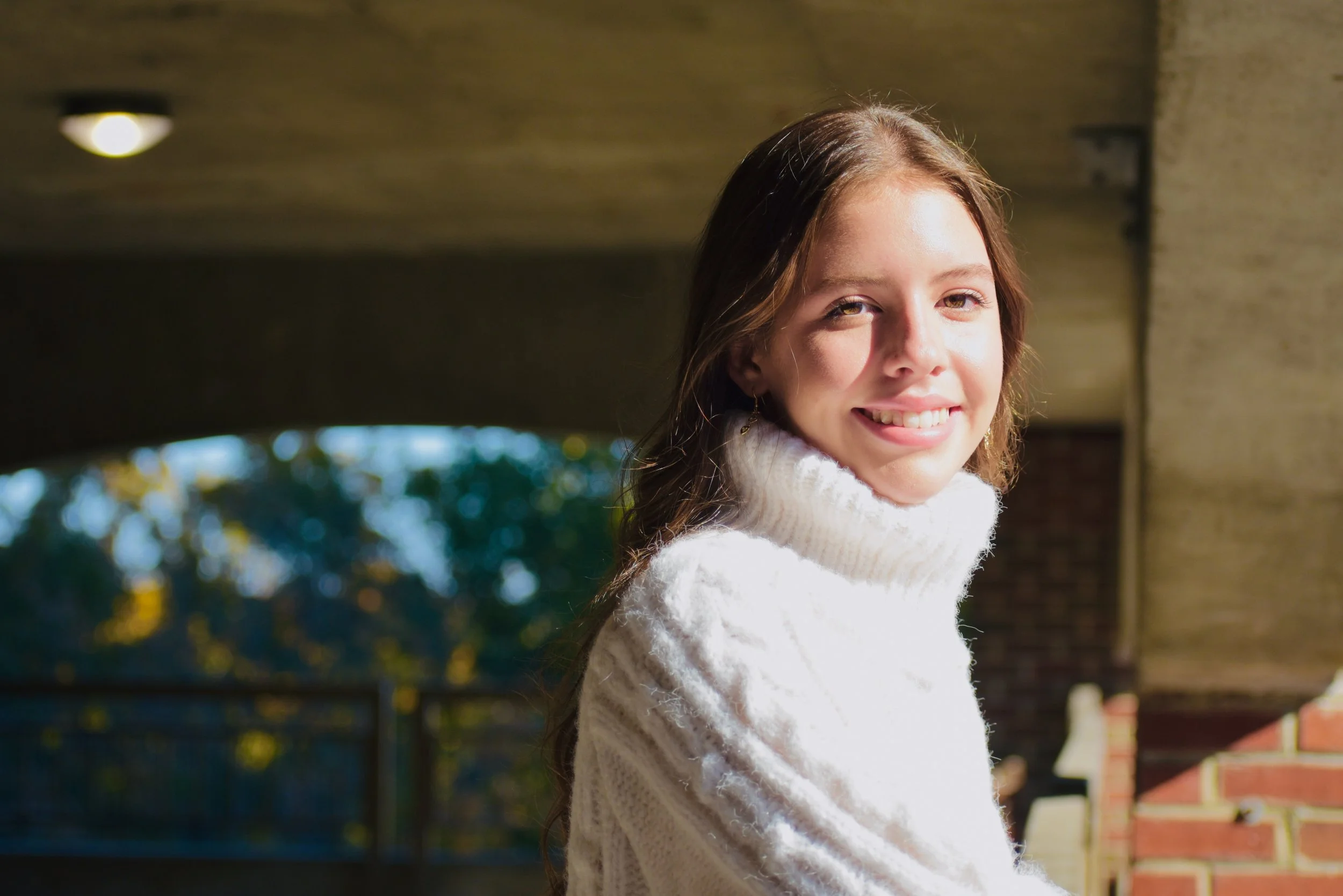 Smiling young woman with long hair wearing a white turtleneck sweater in sunlight outside.