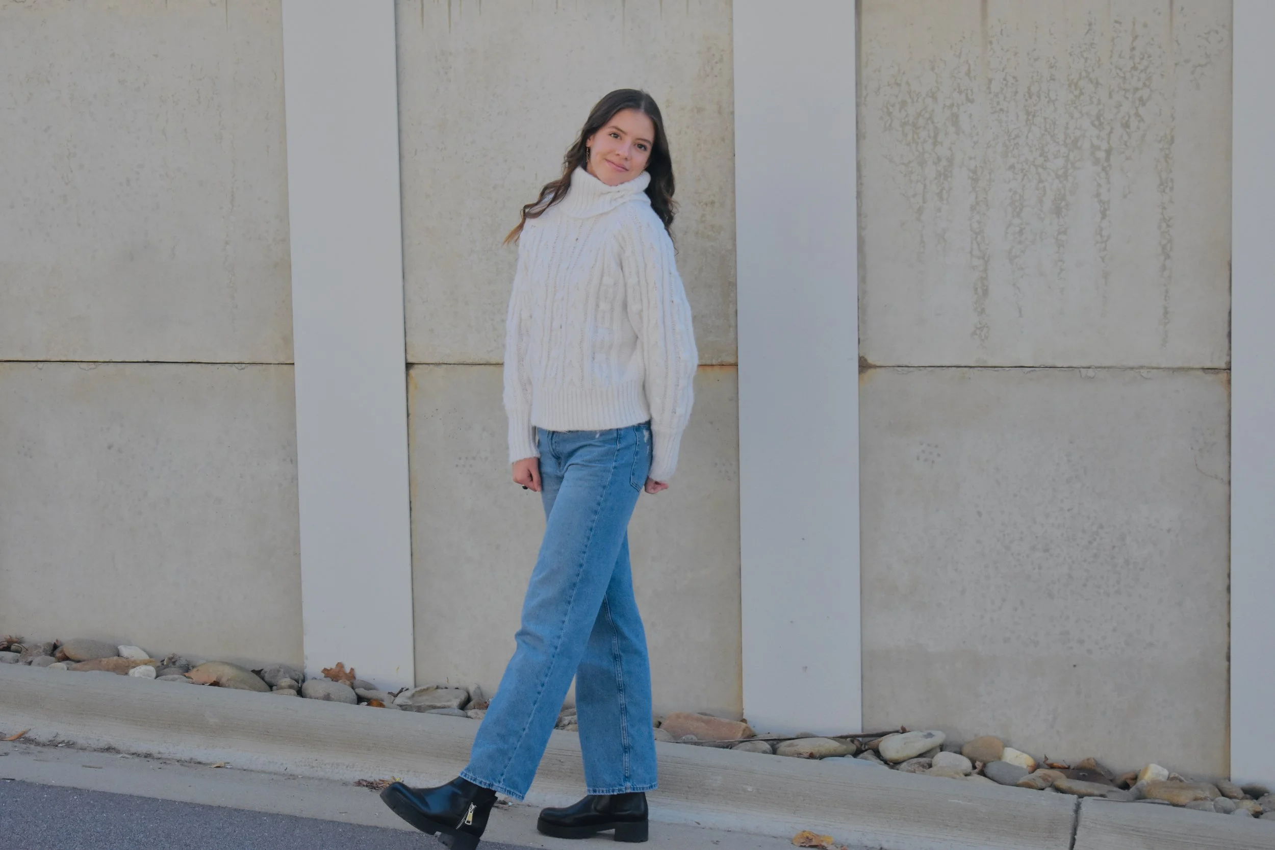 A young woman with long dark hair standing outdoors against a concrete wall, wearing a white cable-knit turtleneck sweater, blue jeans, and black ankle boots, smiling at the camera.
