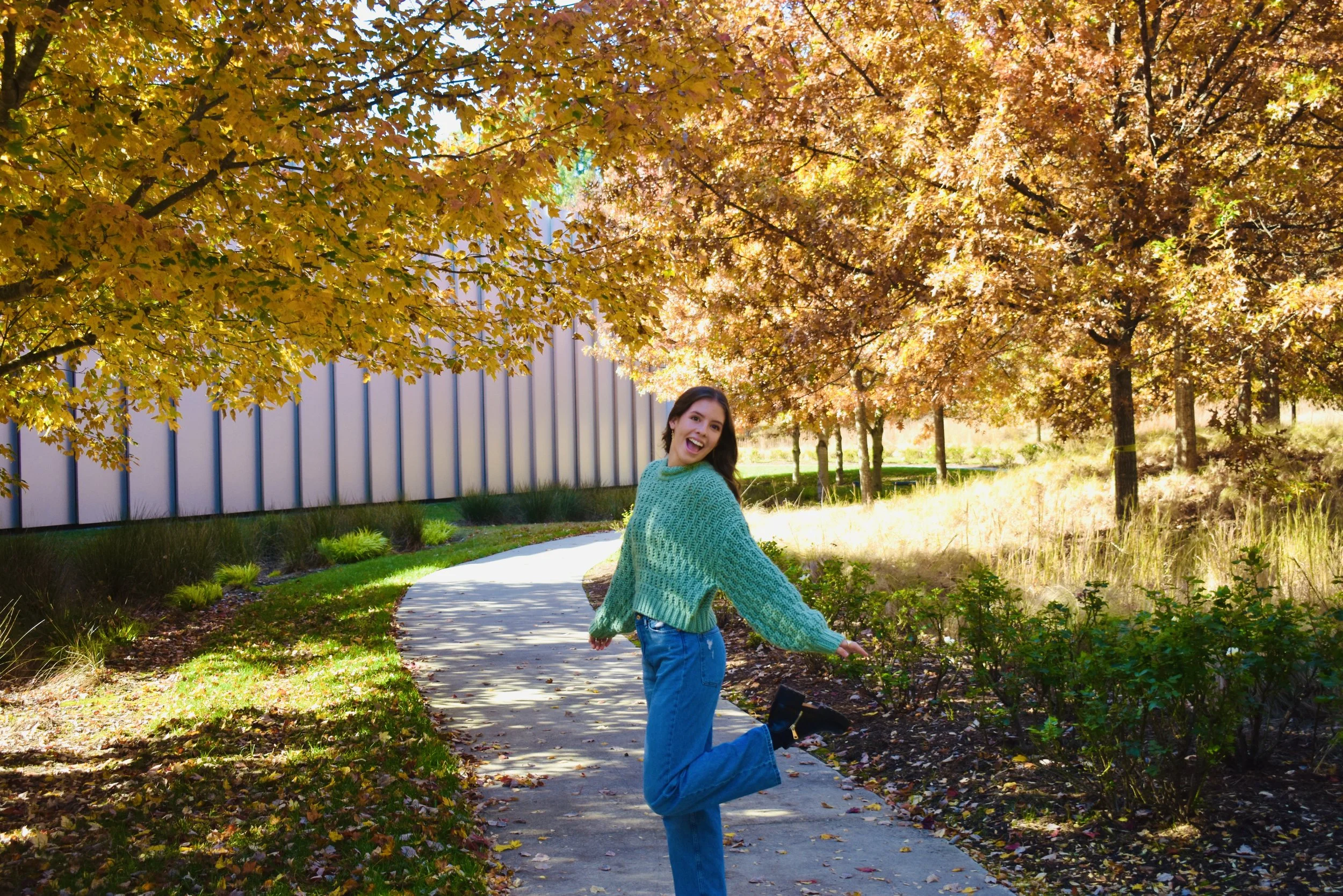 A smiling woman in a teal sweater and blue jeans posing on a winding path surrounded by trees with fall foliage.