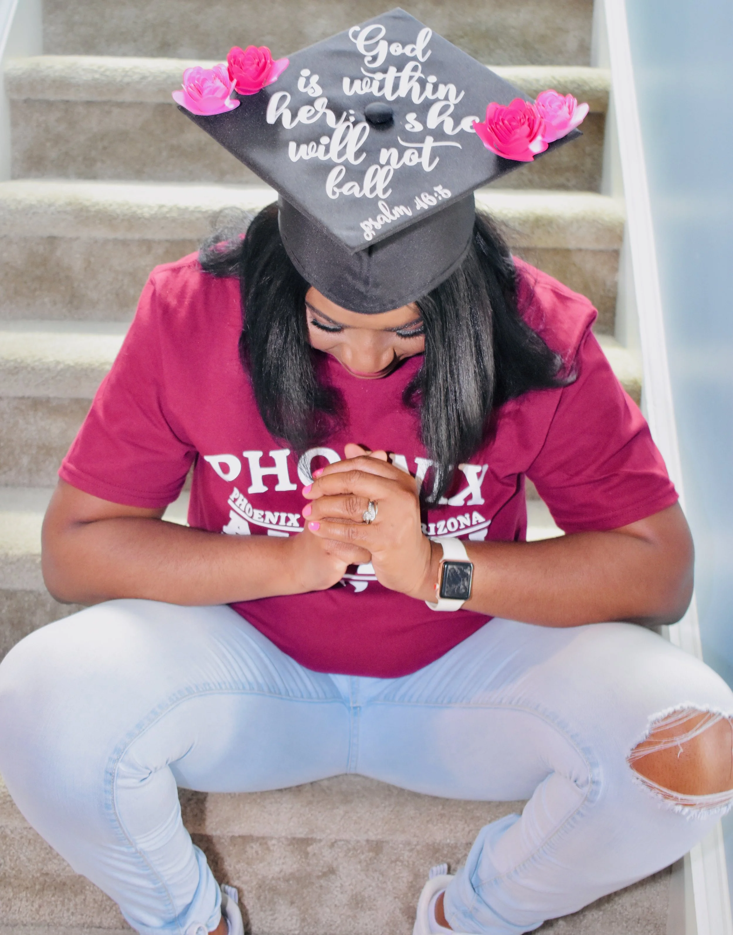 A woman wearing a black graduation cap with pink flowers, a maroon T-shirt, and white ripped jeans sits on stairs with her hands clasped in prayer, looking down.