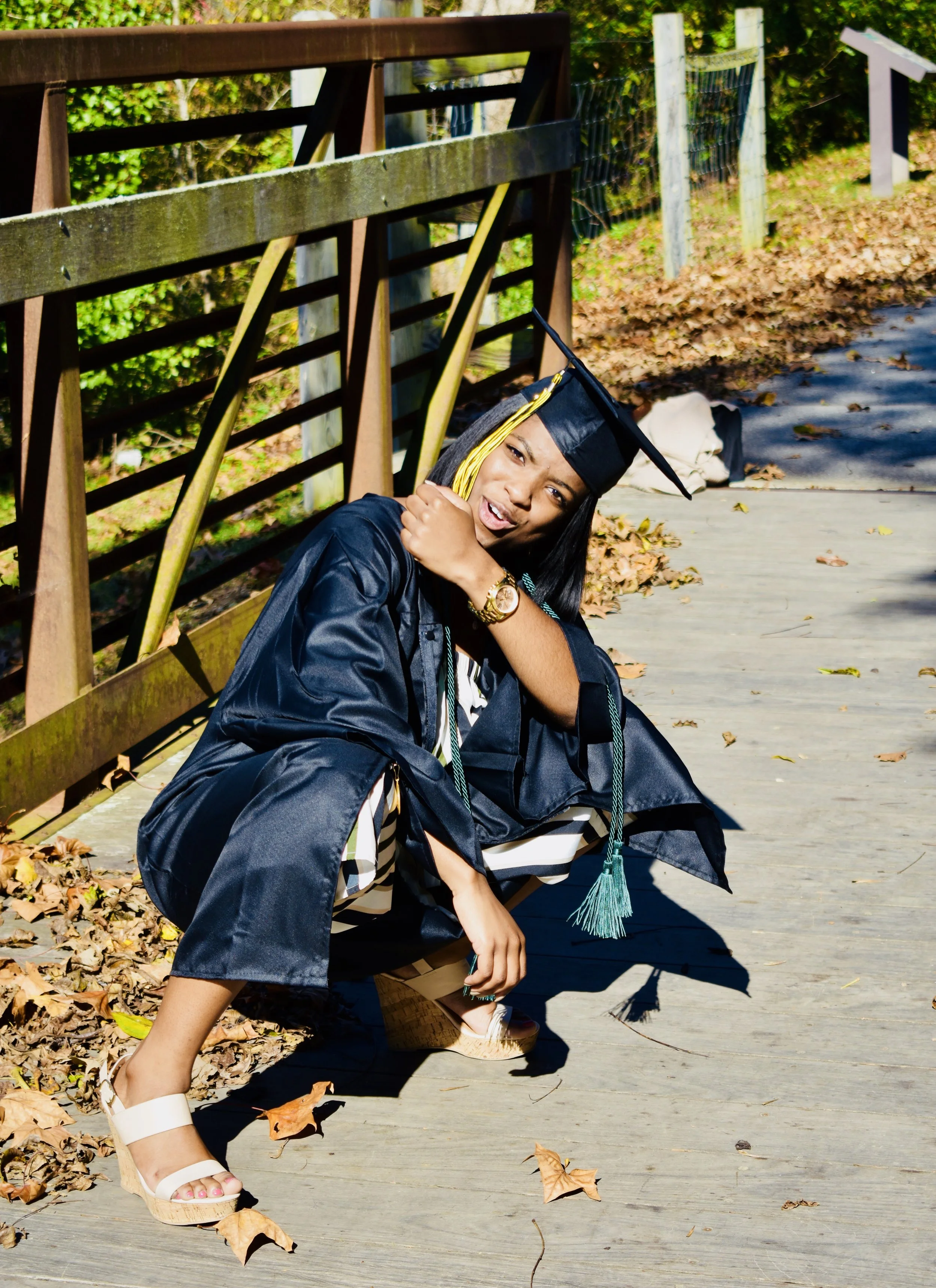 A woman in a black graduation gown and cap with yellow tassel squatting on a wooden outdoor path, gesturing with her hand, and making a playful face.