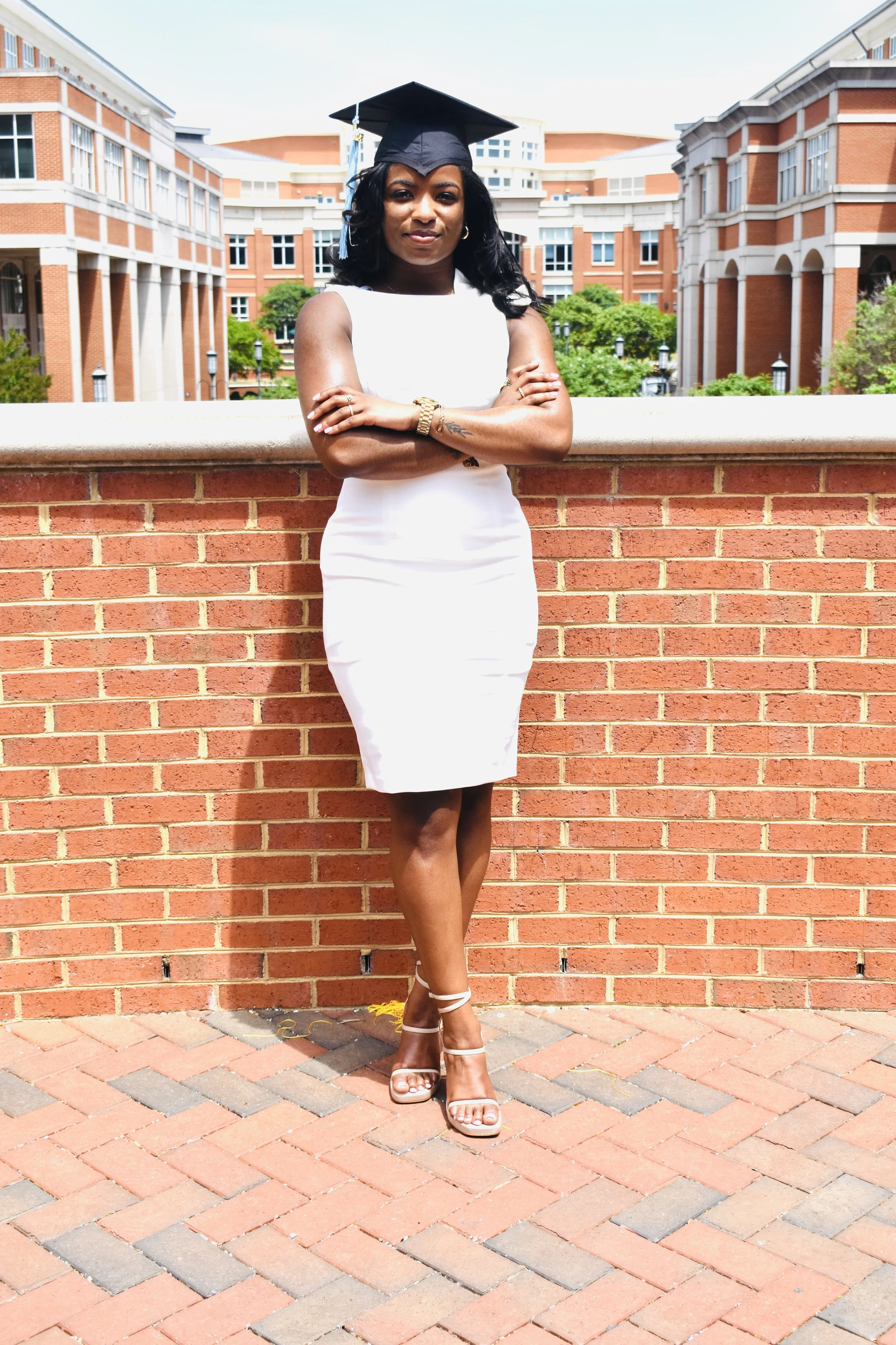 A young woman in a white dress and high heels stands outdoors in front of a brick wall, wearing a black graduation cap, with arms crossed and a confident expression.