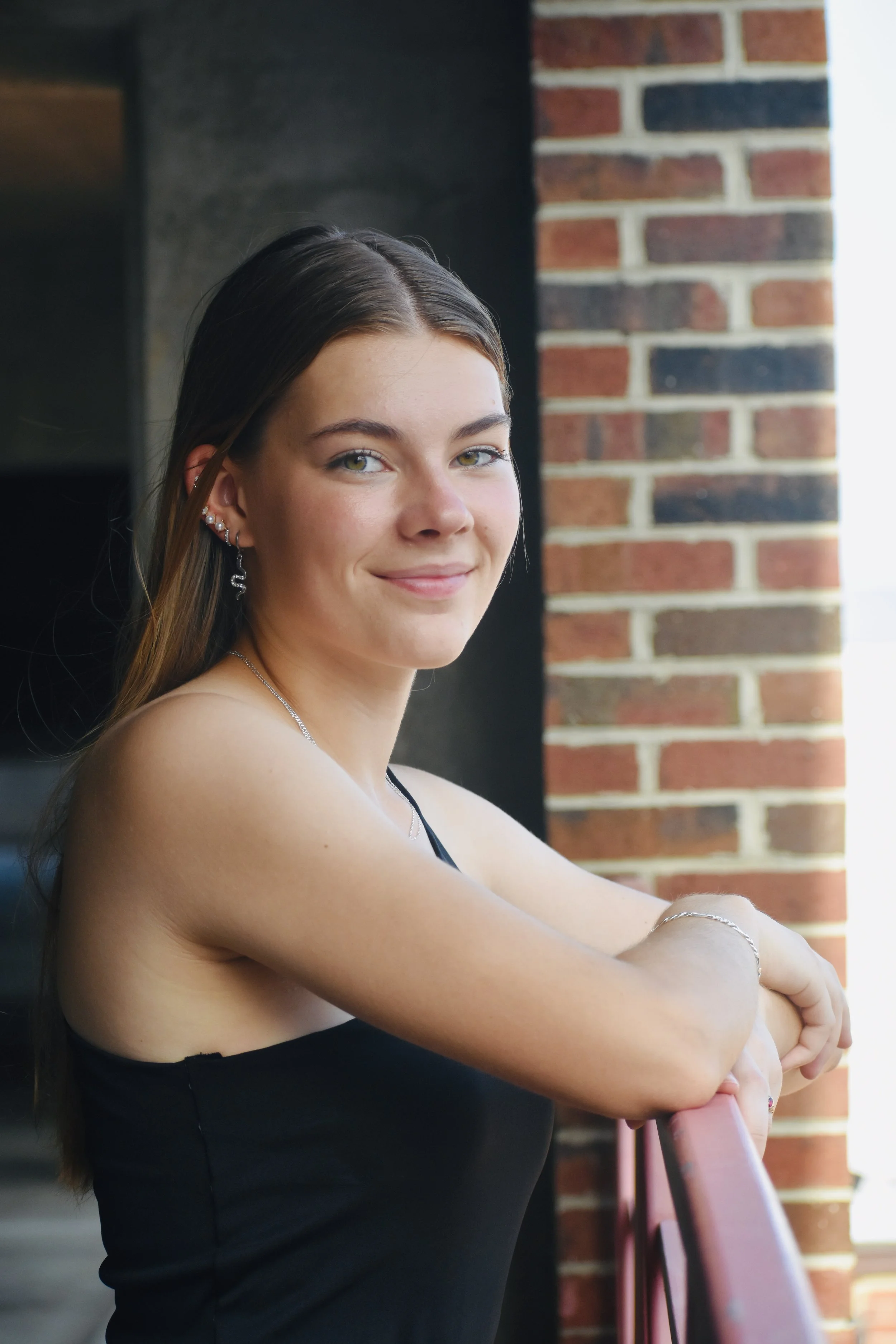 Young woman with long brown hair and silver jewelry standing near a brick wall, smiling at the camera.