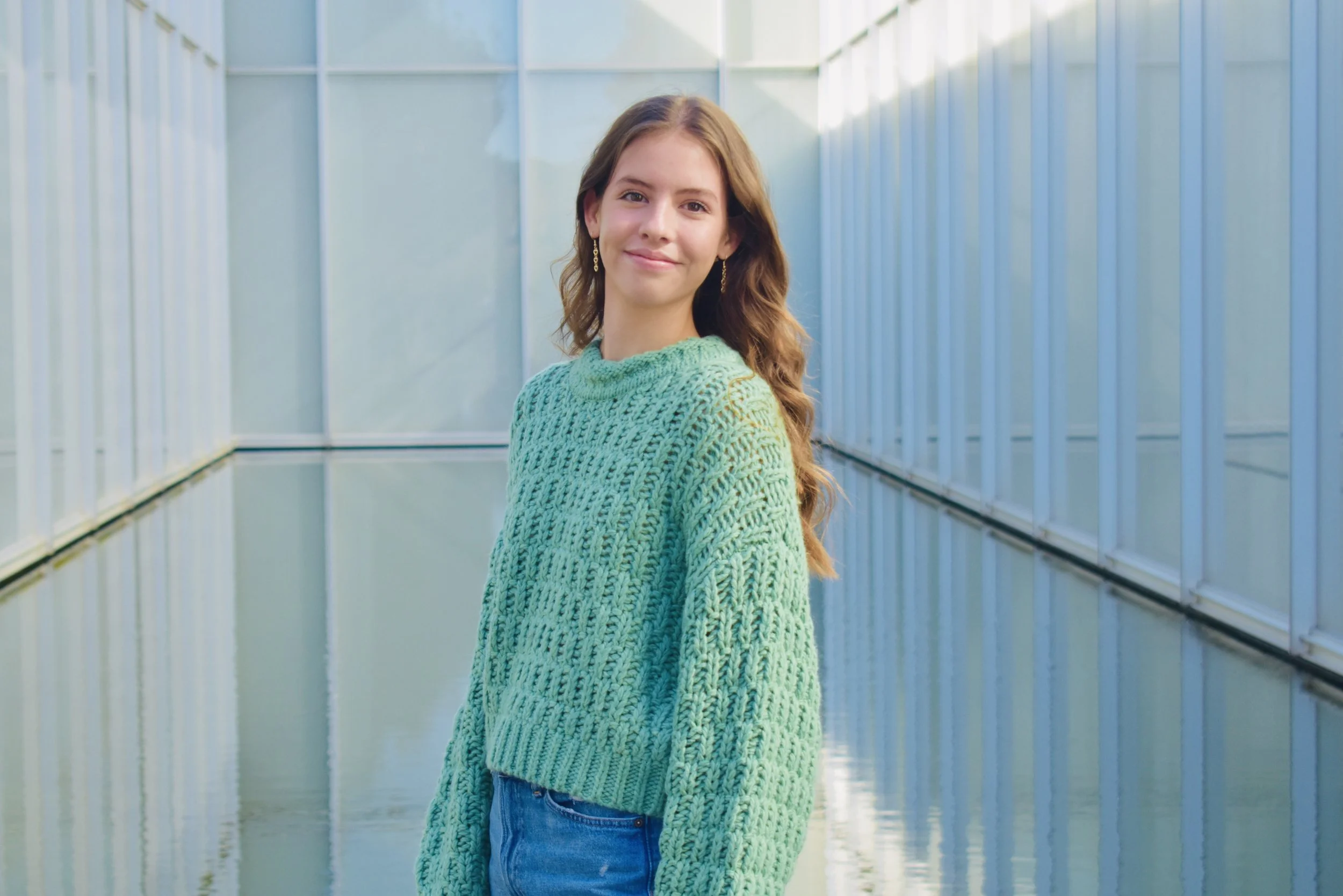 A young woman with long, wavy brown hair smiling standing in a glass-enclosed corridor with blue metal framing, wearing a chunky green sweater and blue jeans.