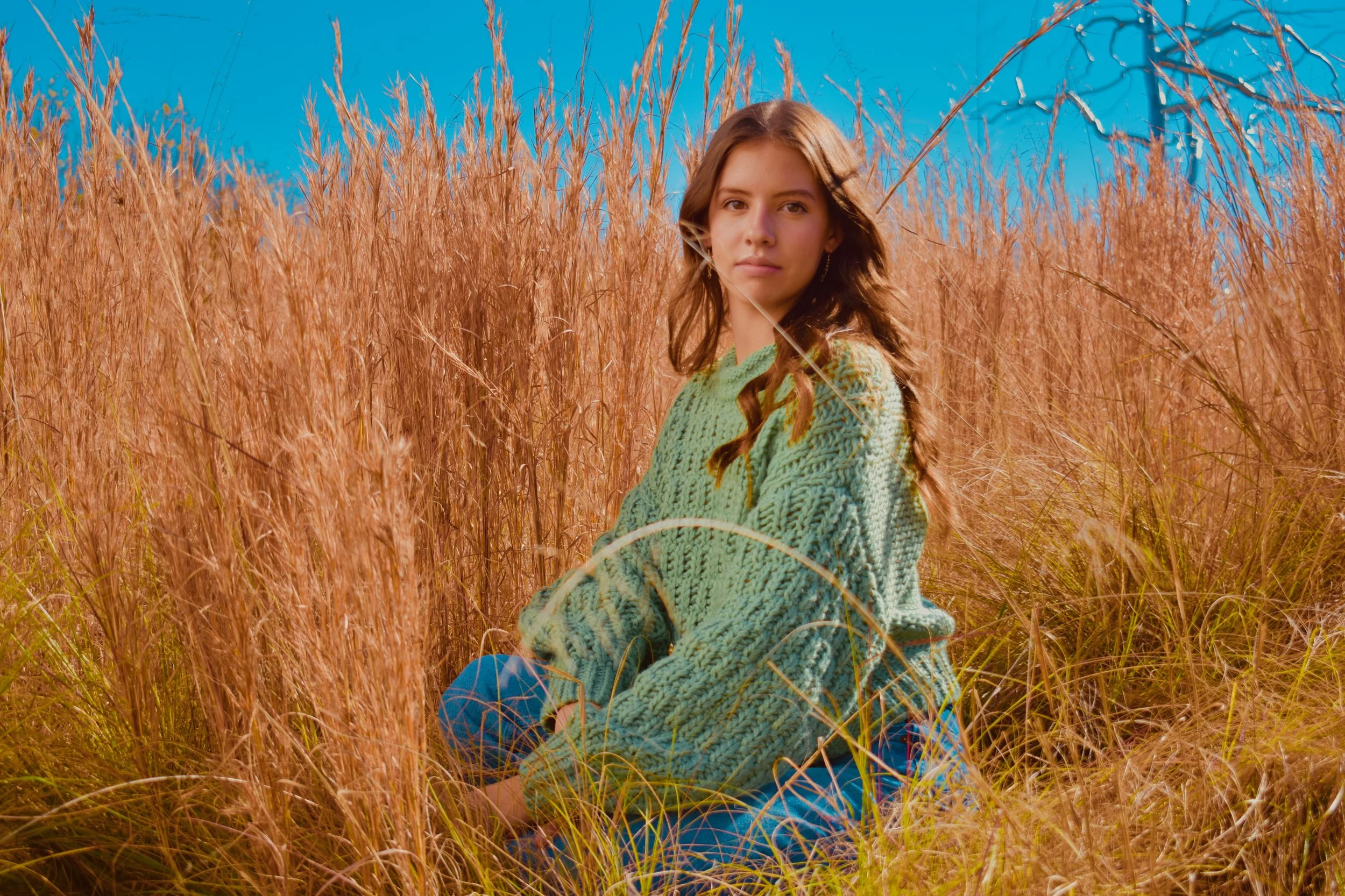 A woman sitting in a field of tall golden grass under a blue sky, wearing a green knit sweater.