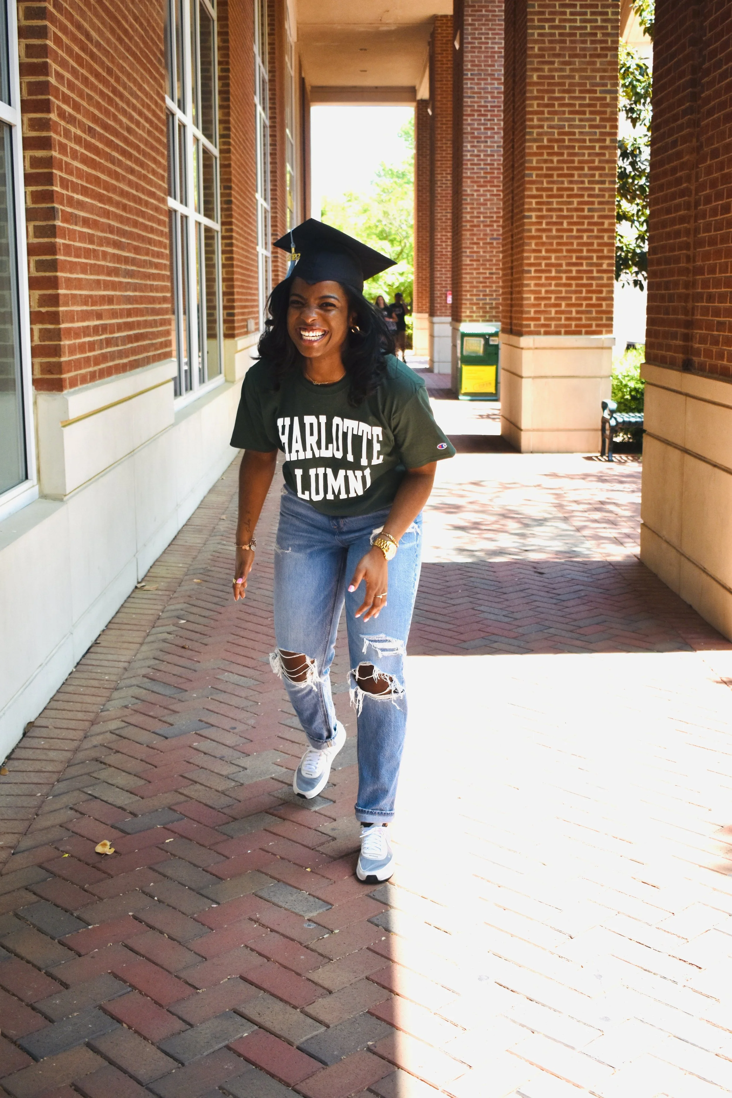 A young woman in a Charlotte Lumni graduation cap and green t-shirt with ripped jeans, smiling and walking outdoors on a brick sidewalk.