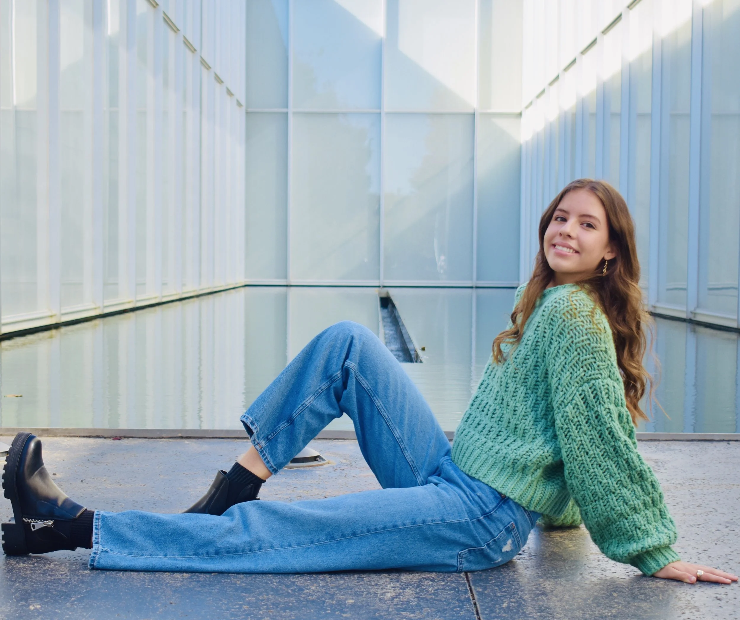 Young woman with long wavy hair, wearing a green knitted sweater and blue jeans, sitting on the ground in front of a modern glass building, smiling at the camera.