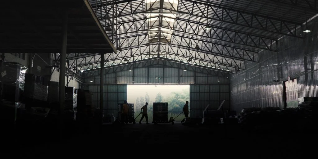 Silhouetted workers moving pallets in a large warehouse with high metal roofing and stacks of products, light streaming through an open door at the far end.