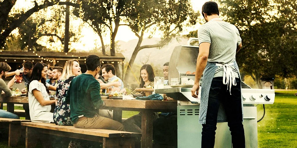 Group of people enjoying a barbecue in a grassy outdoor setting with picnic tables, trees, and string lights.