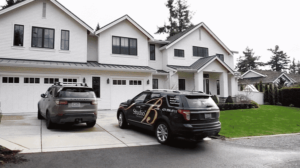 Large white two-story house with three garage doors, two parked SUVs in the driveway, and a green lawn with shrubs.