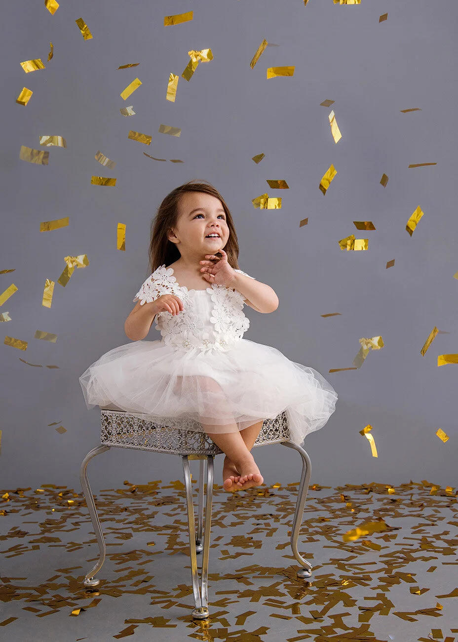 A young girl wearing a white dress sitting on a metal stool, surrounded by falling gold confetti and a layer of confetti on the floor, against a gray background.