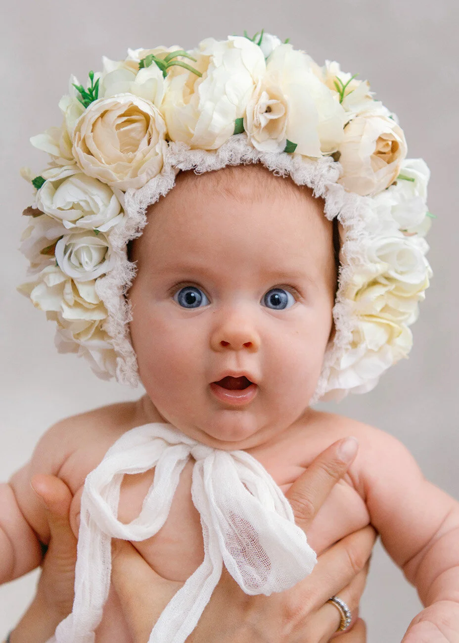 Baby with big blue eyes wearing a floral bonnet made of white and cream roses, holding a white ribbon tied around their neck.