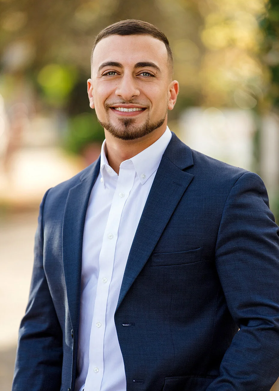 A young man with short brown hair, a trimmed beard, wearing a navy suit and white dress shirt, smiling outdoors with a blurred green and yellow background.