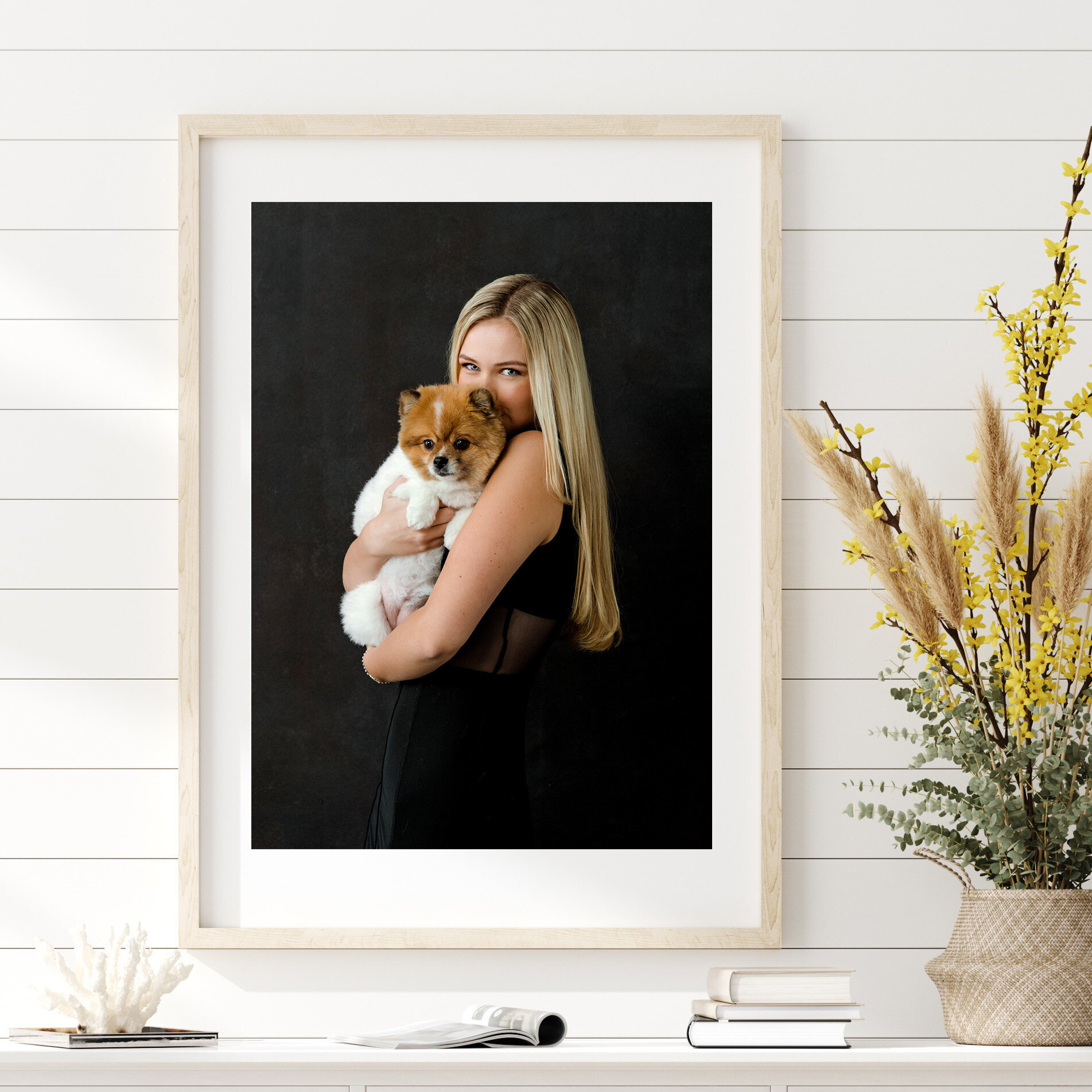 Framed portrait of a woman with long blonde hair holding a small, fluffy puppy against a dark background, decorated with a vase of yellow flowers and pampas grass on a white wall.